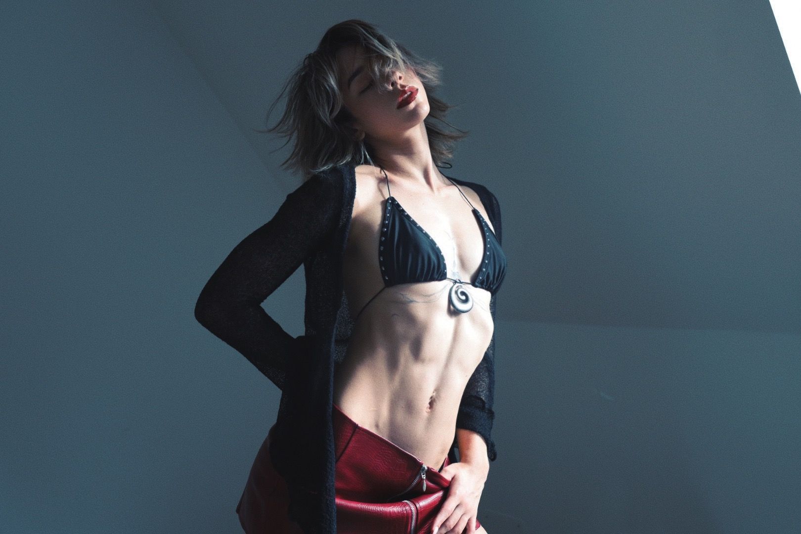 Woman in red pants, black bikini top, and jacket poses, leaning back with eyes closed.