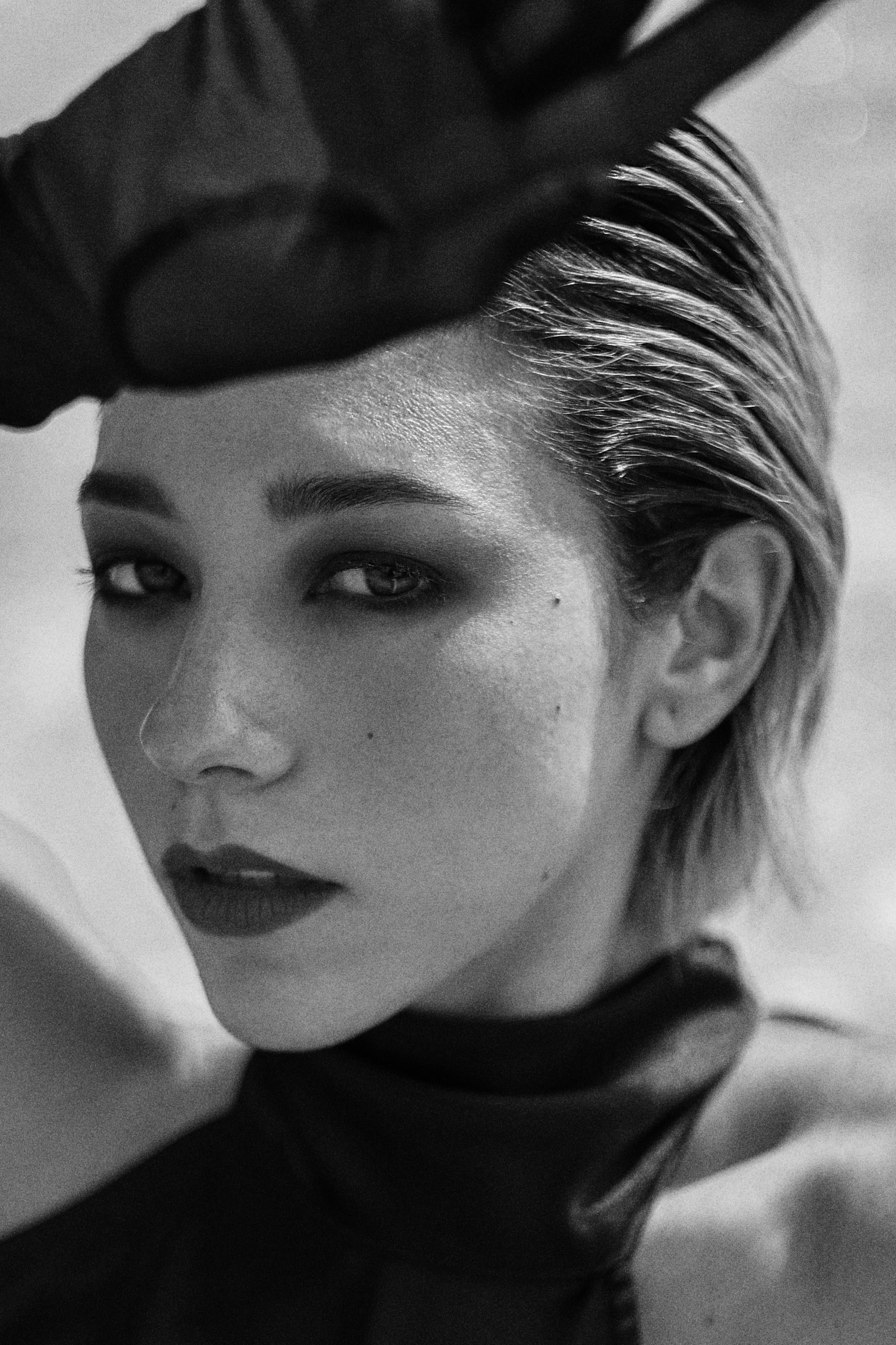 Woman with dark eye makeup and red lipstick, looking towards the camera, gloved hand above her head.