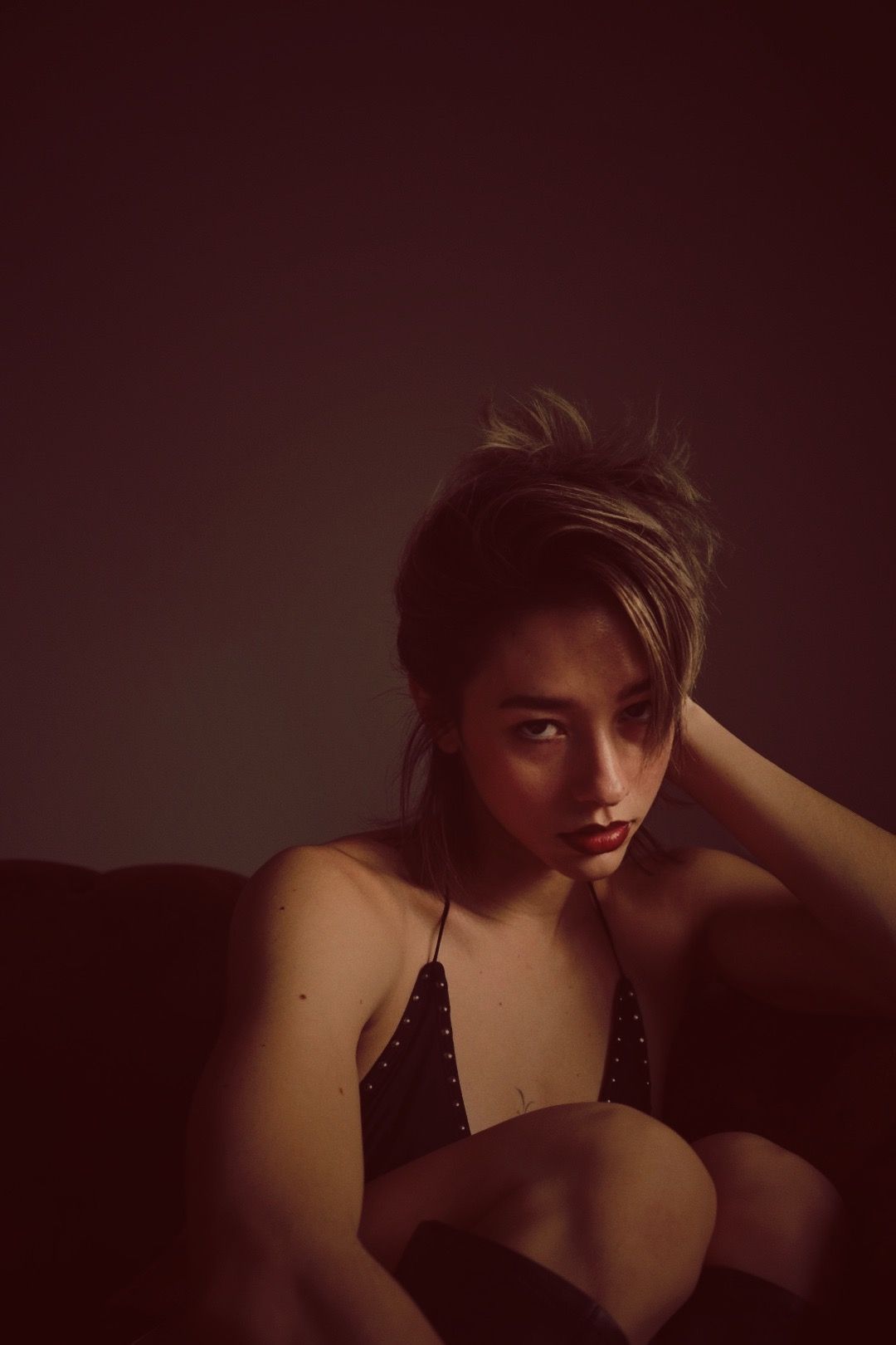 Woman in black top, leaning, looking forward with red lipstick, dark background.