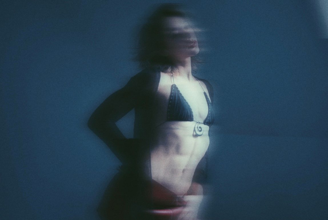 Blurred figure in a bikini, lit by a side light, against a blue wall.