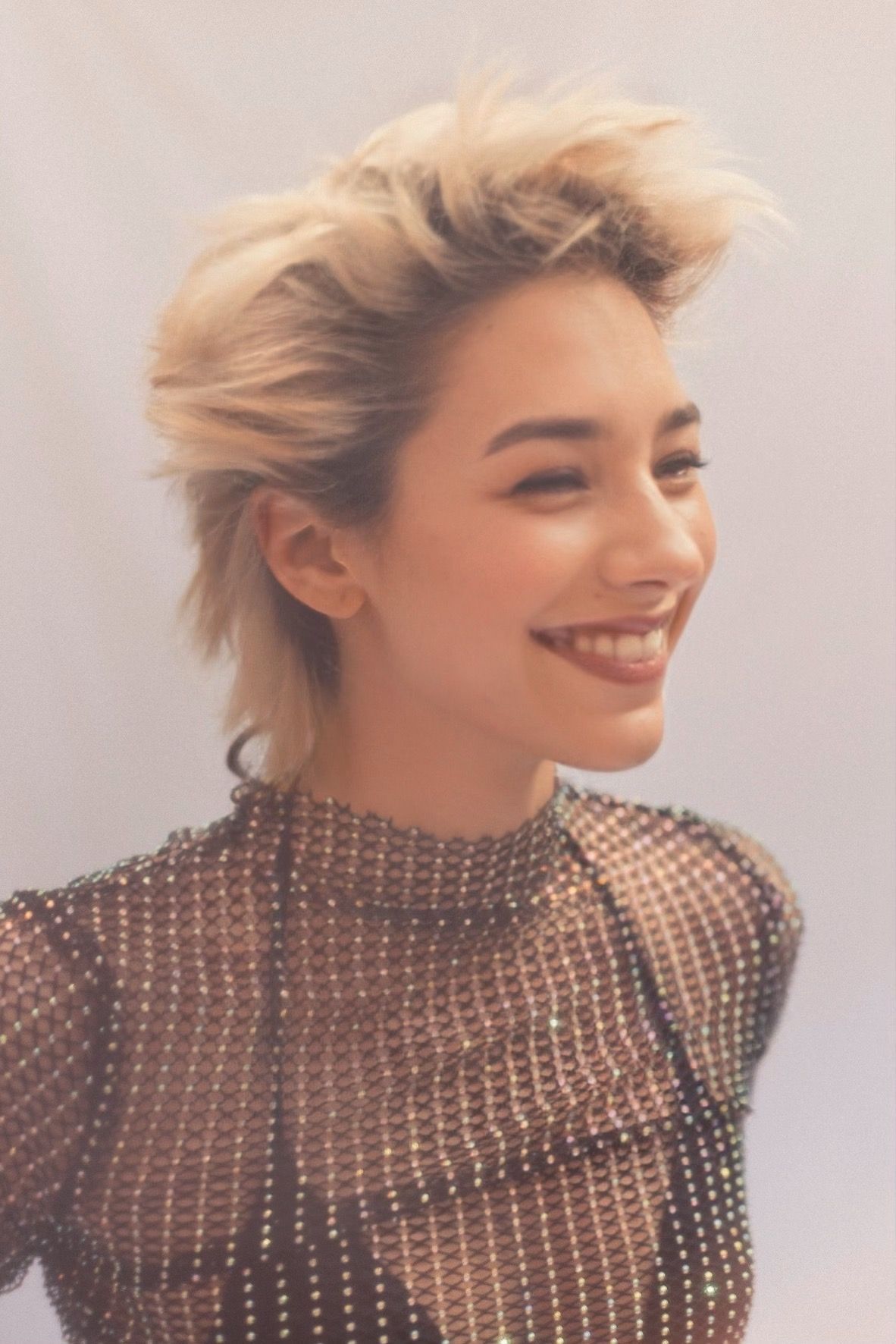 Woman with short, spiky blonde hair, smiling. Wearing a sheer, jewel-studded top.