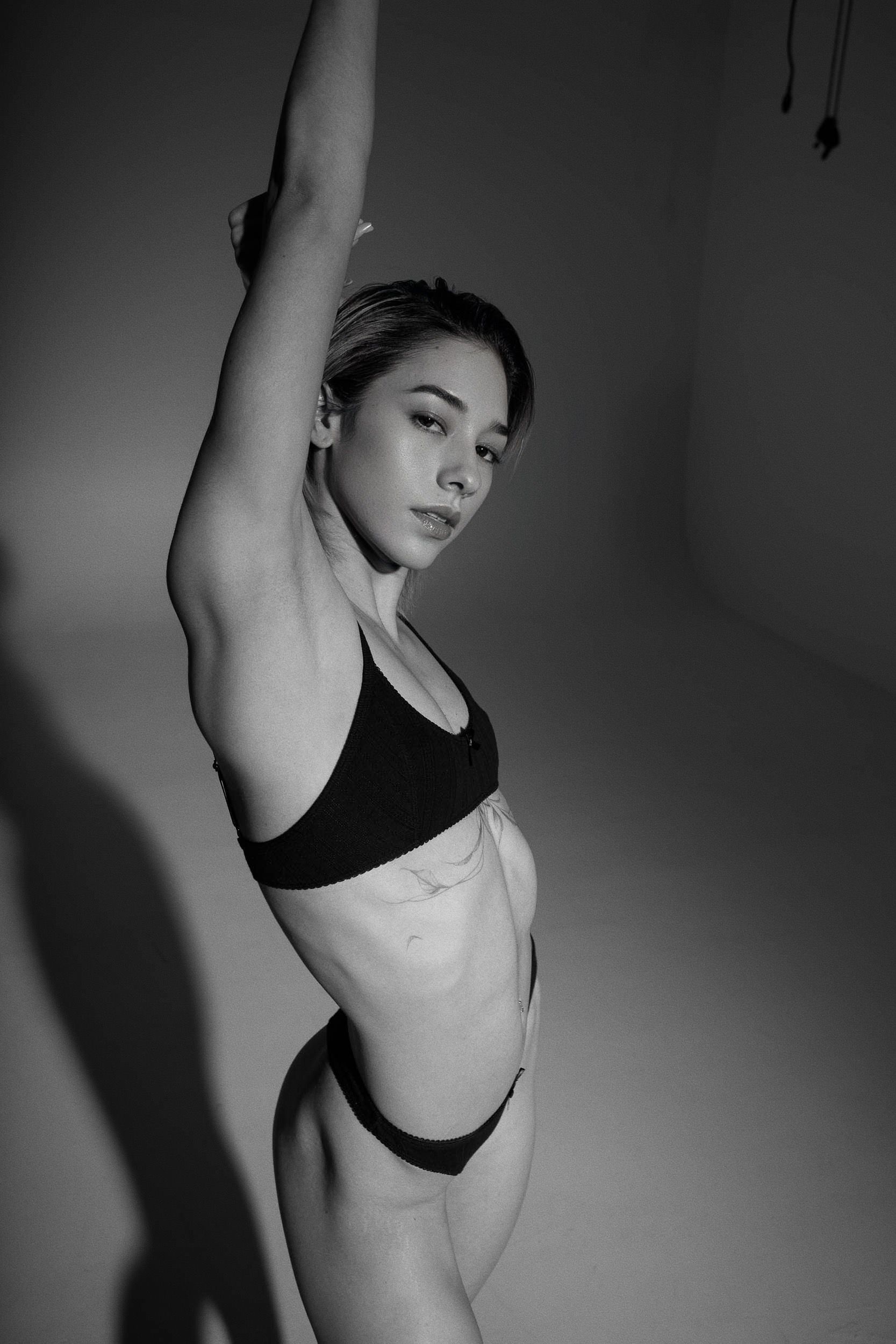 Woman in black lingerie, arm raised, posing against a neutral wall.