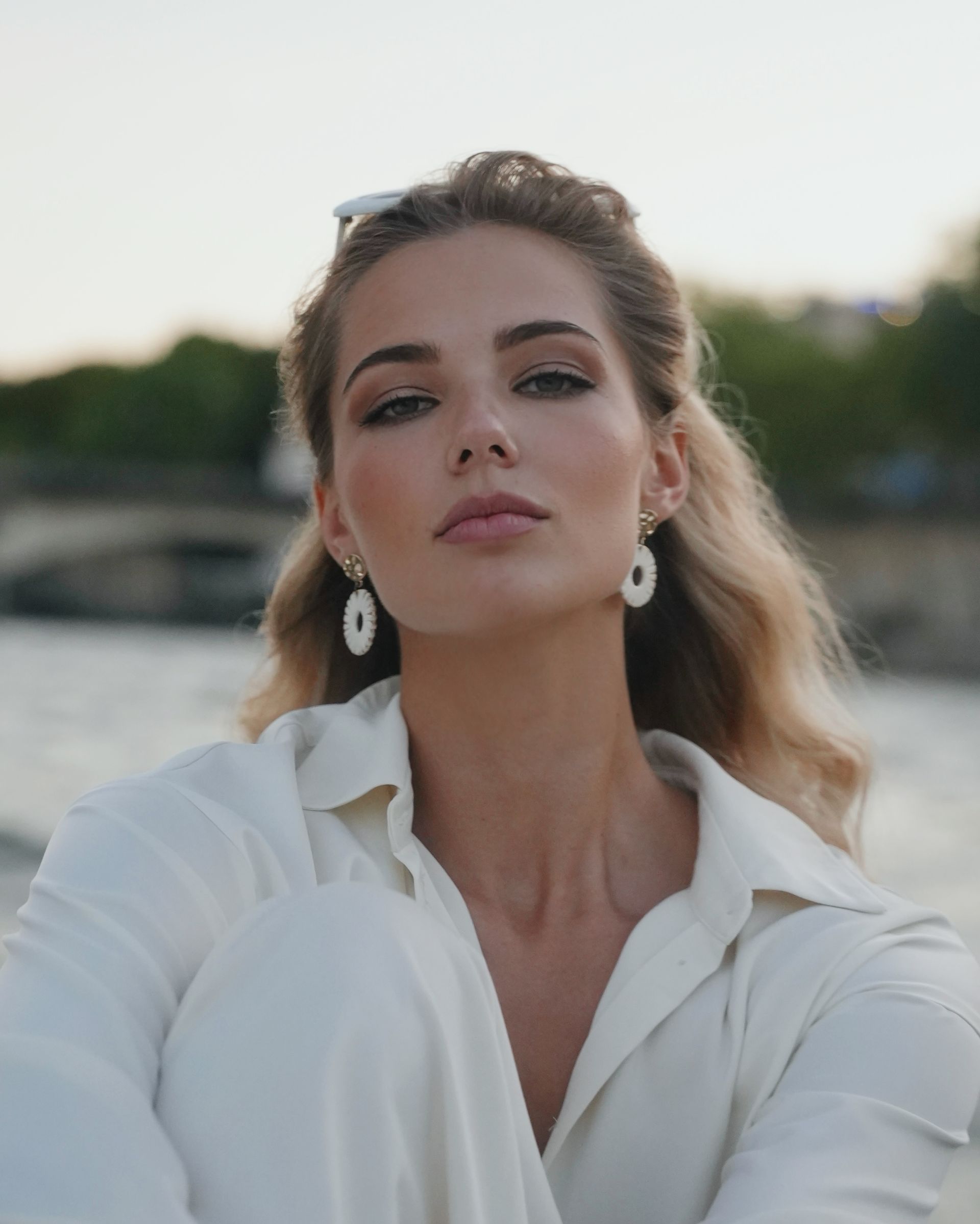 Woman with blonde hair wearing white outfit and earrings, looking at camera outdoors.
