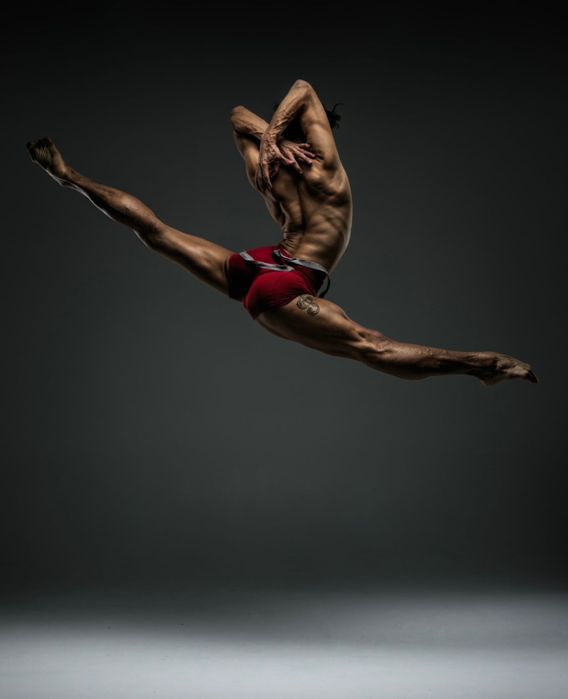 Male dancer in mid-air split, arms overhead, wearing red shorts. Dark background.