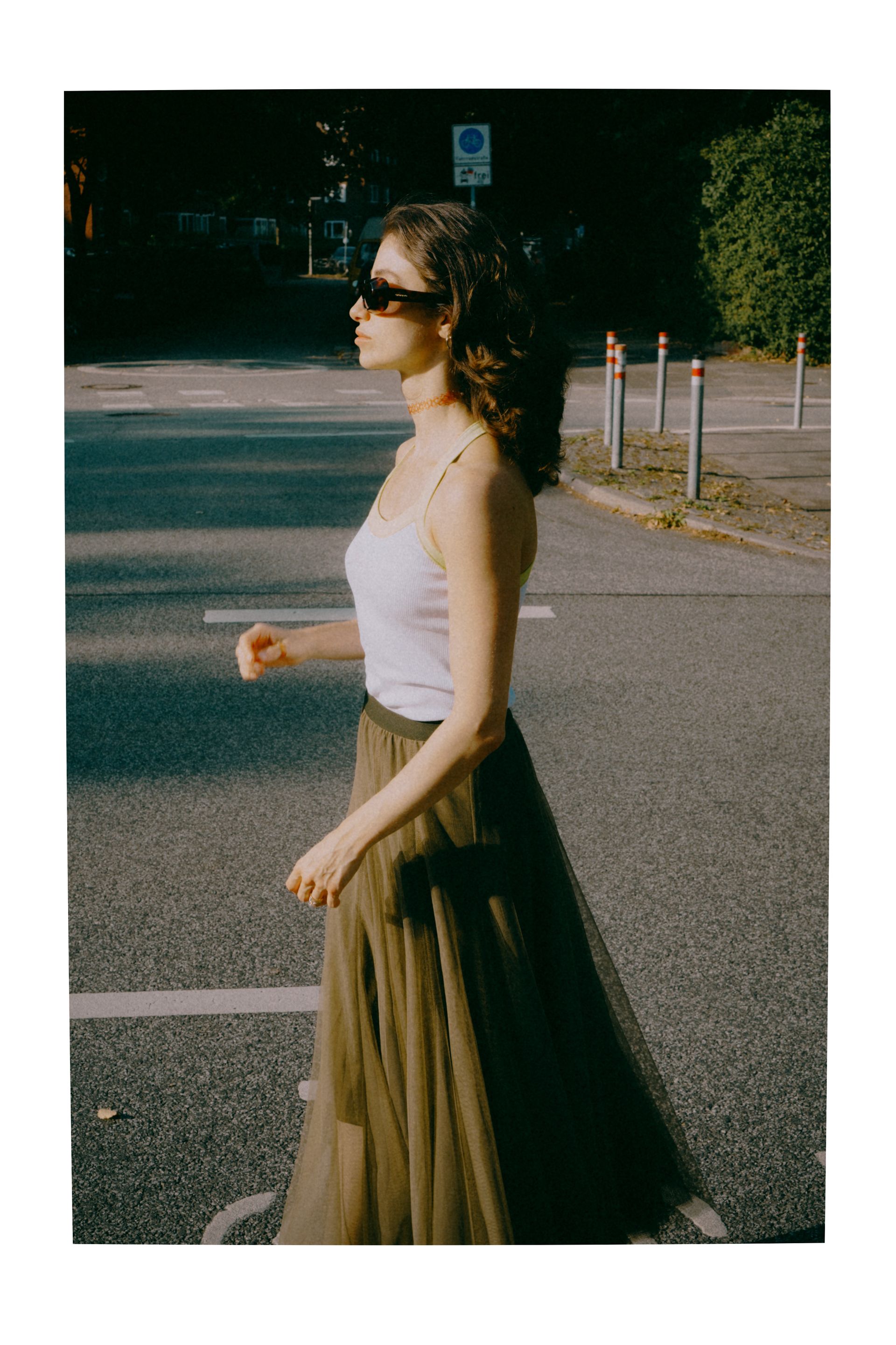 Woman in sunglasses walks across a street in a light purple top and dark green skirt.