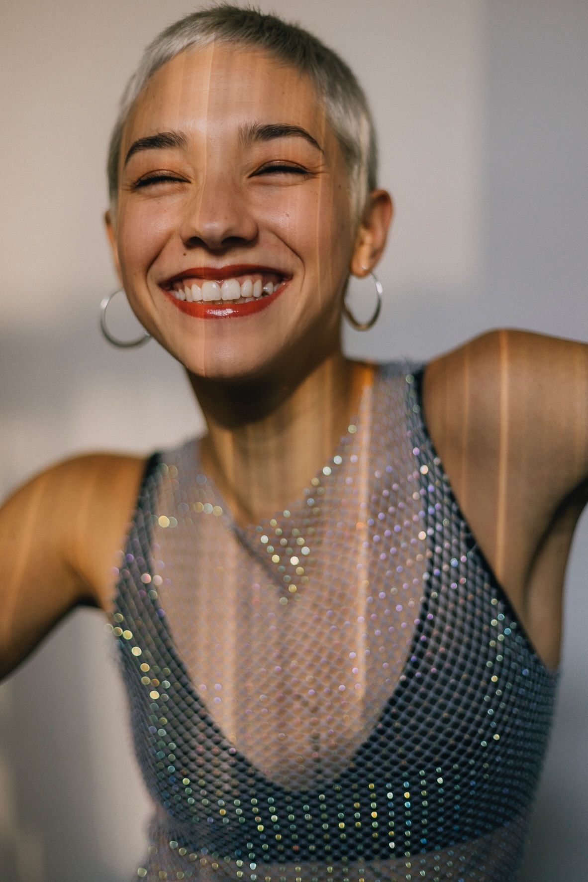 Woman with a short, light-colored haircut, wearing a shimmering top, smiling broadly with hoop earrings.