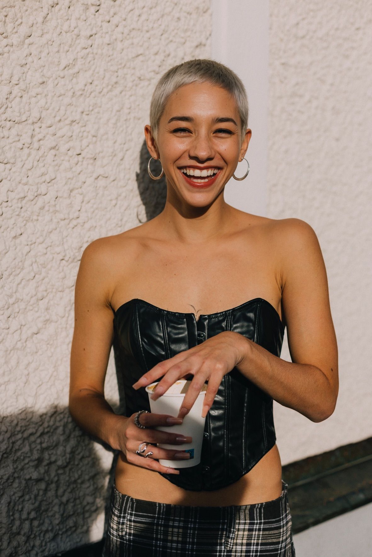 Woman with short, light hair smiles, holding cup, wearing black corset and plaid pants outside.