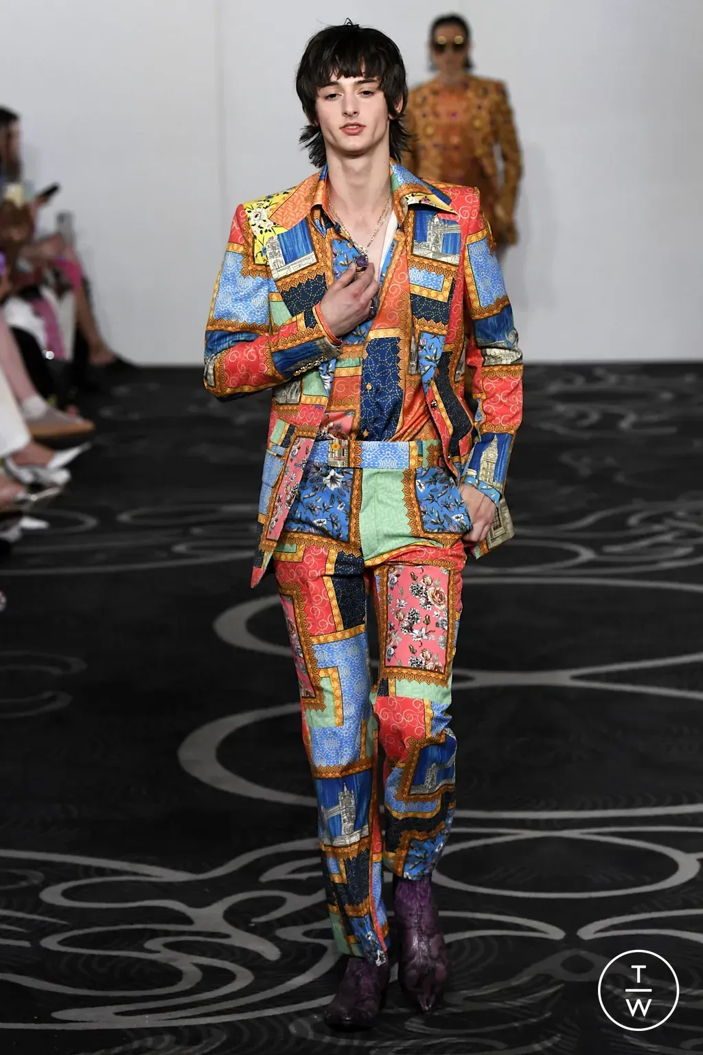 Man walking runway in patchwork suit, purple boots. Floral carpet, white backdrop.