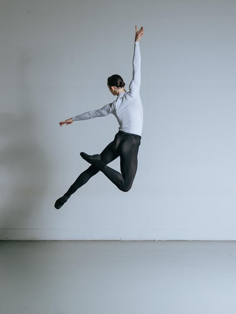 Male dancer in mid-air, arms and legs extended, wearing gray top and dark tights, set against a plain wall.