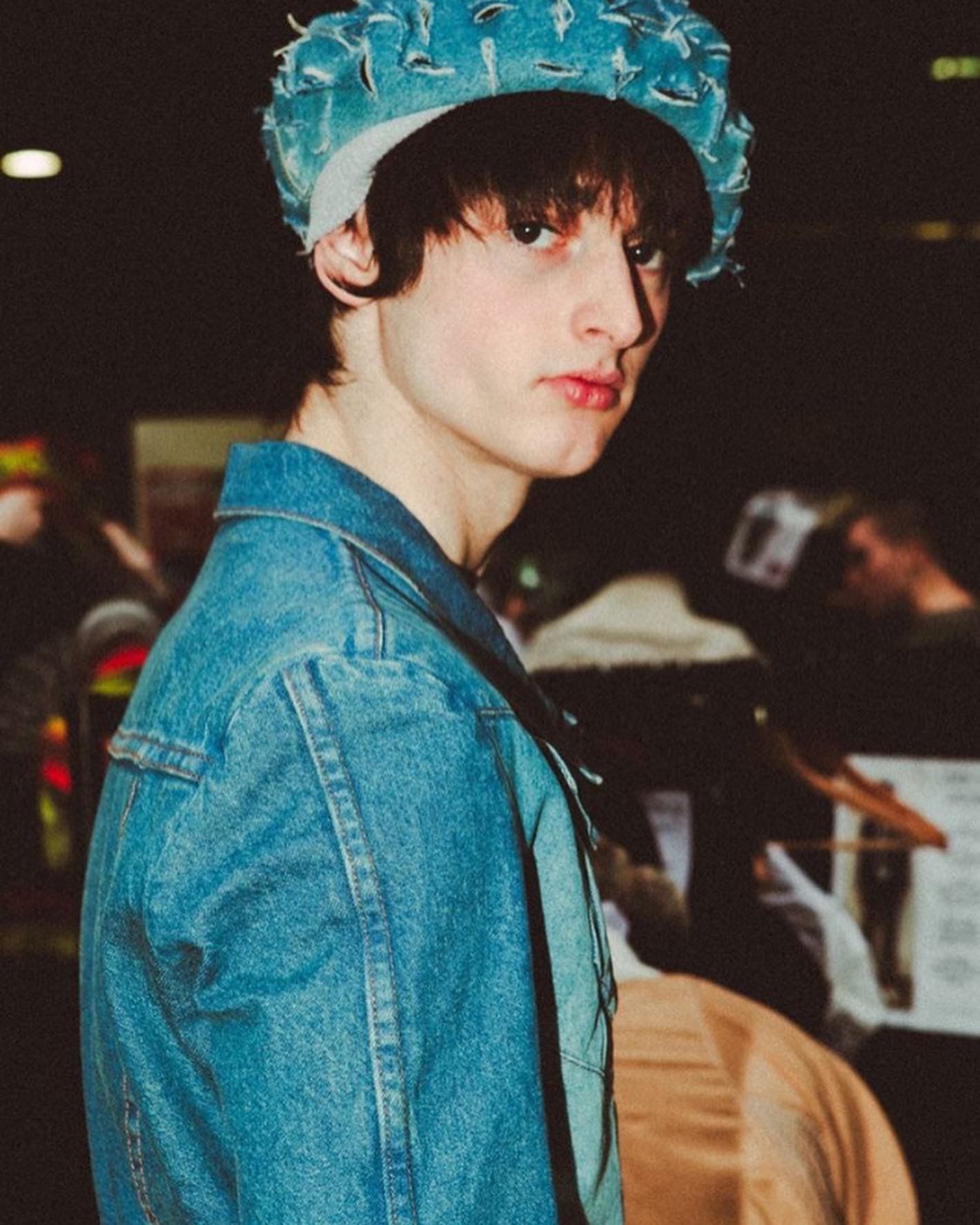 Person in a denim jacket and blue hat, looking at the camera in a crowded area.