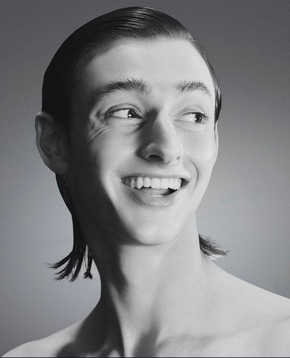 Smiling person with slicked-back hair and a ponytail, looking to the side. Black and white.