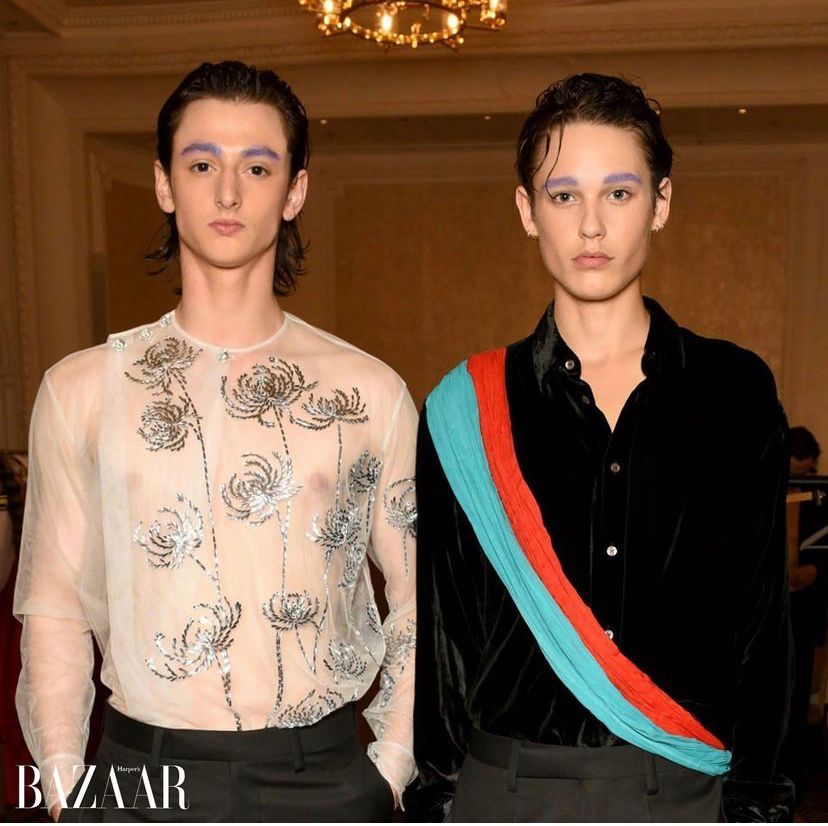 Two men pose; one in sheer shirt with flower embellishments, the other in black shirt with sash; both have purple eyebrows.