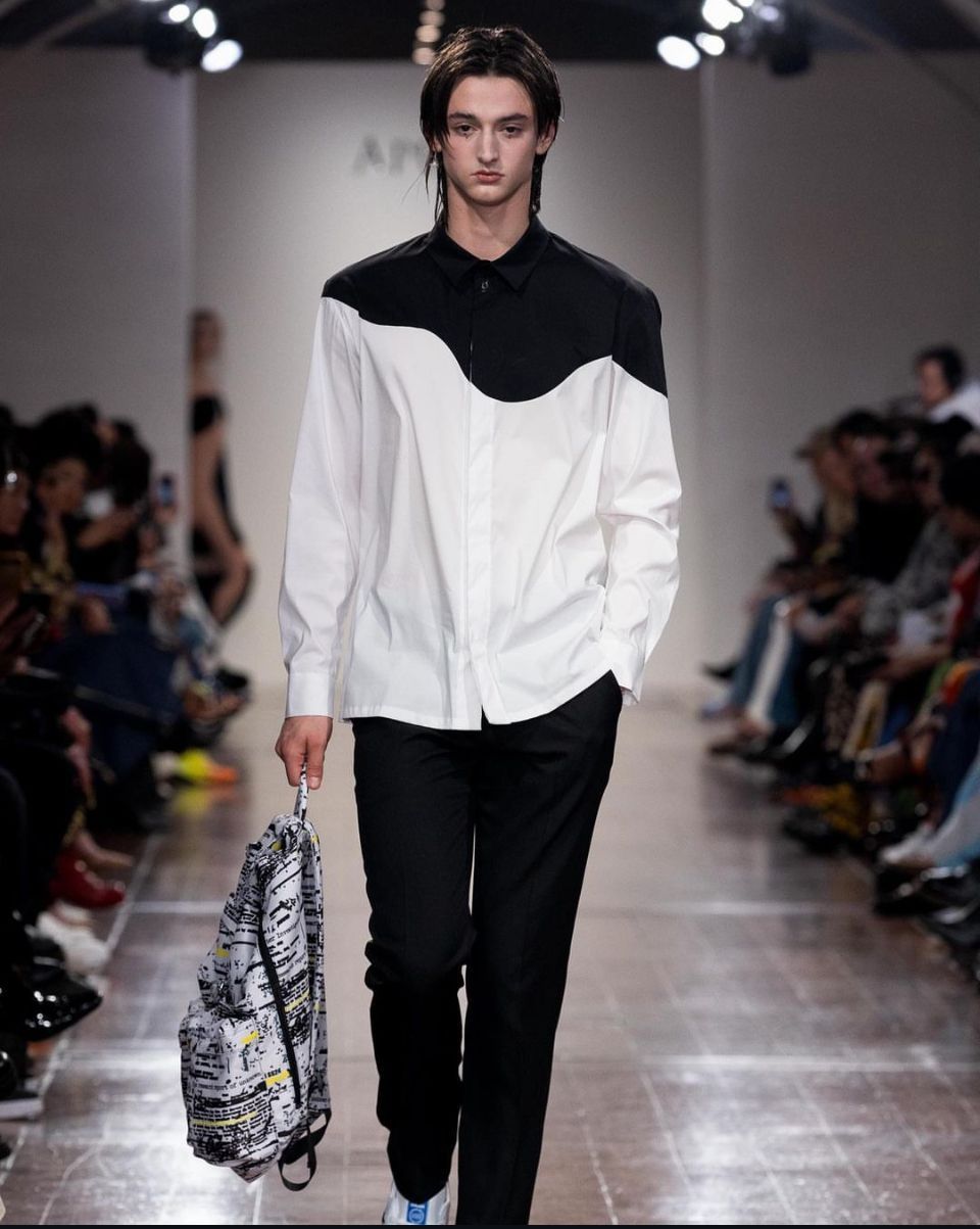 Man in white and black shirt, black pants, walking runway carrying backpack; audience in background.