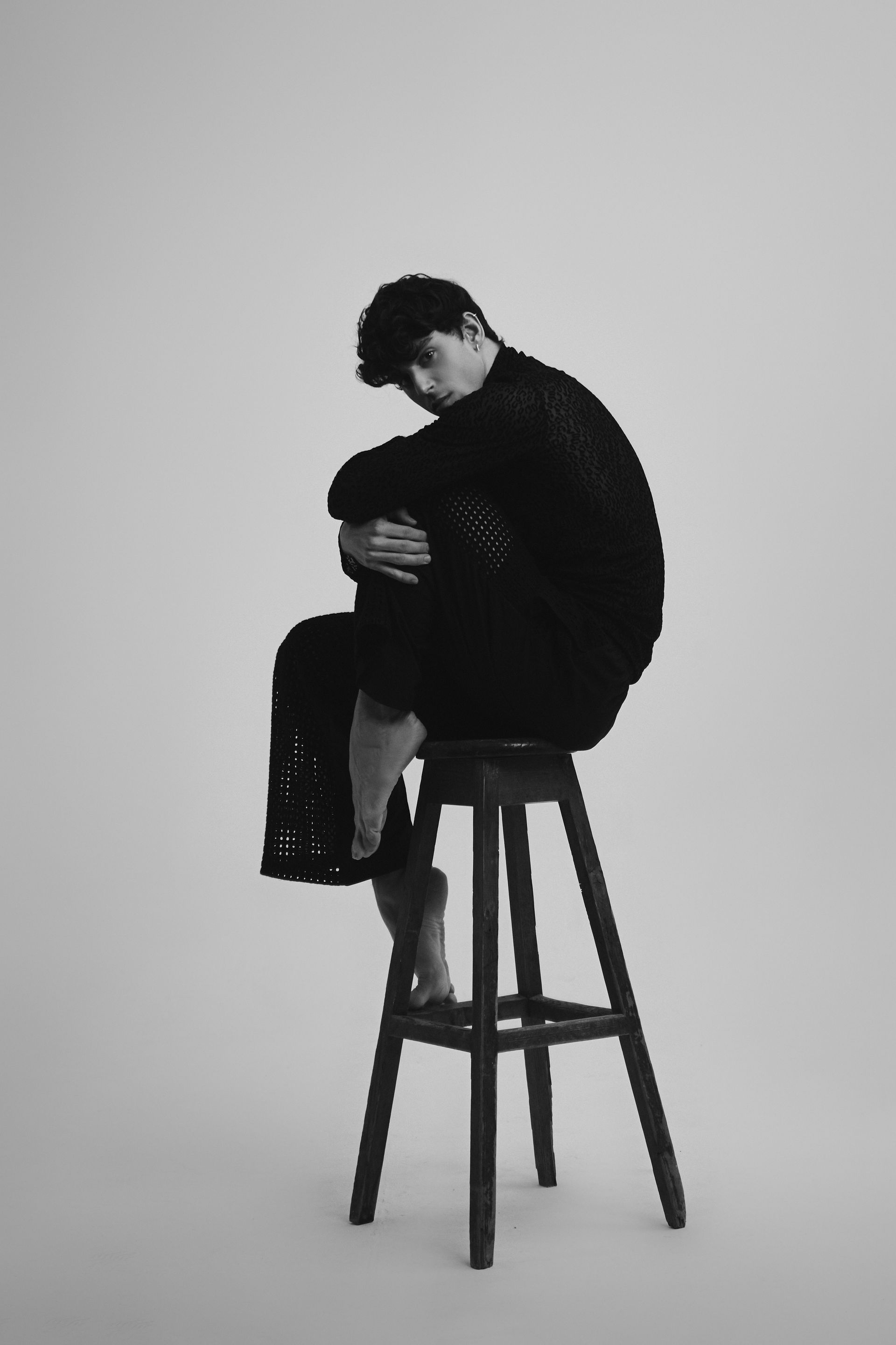 Person sits curled on a stool, wearing black clothing, looking down. Bare feet, white background.