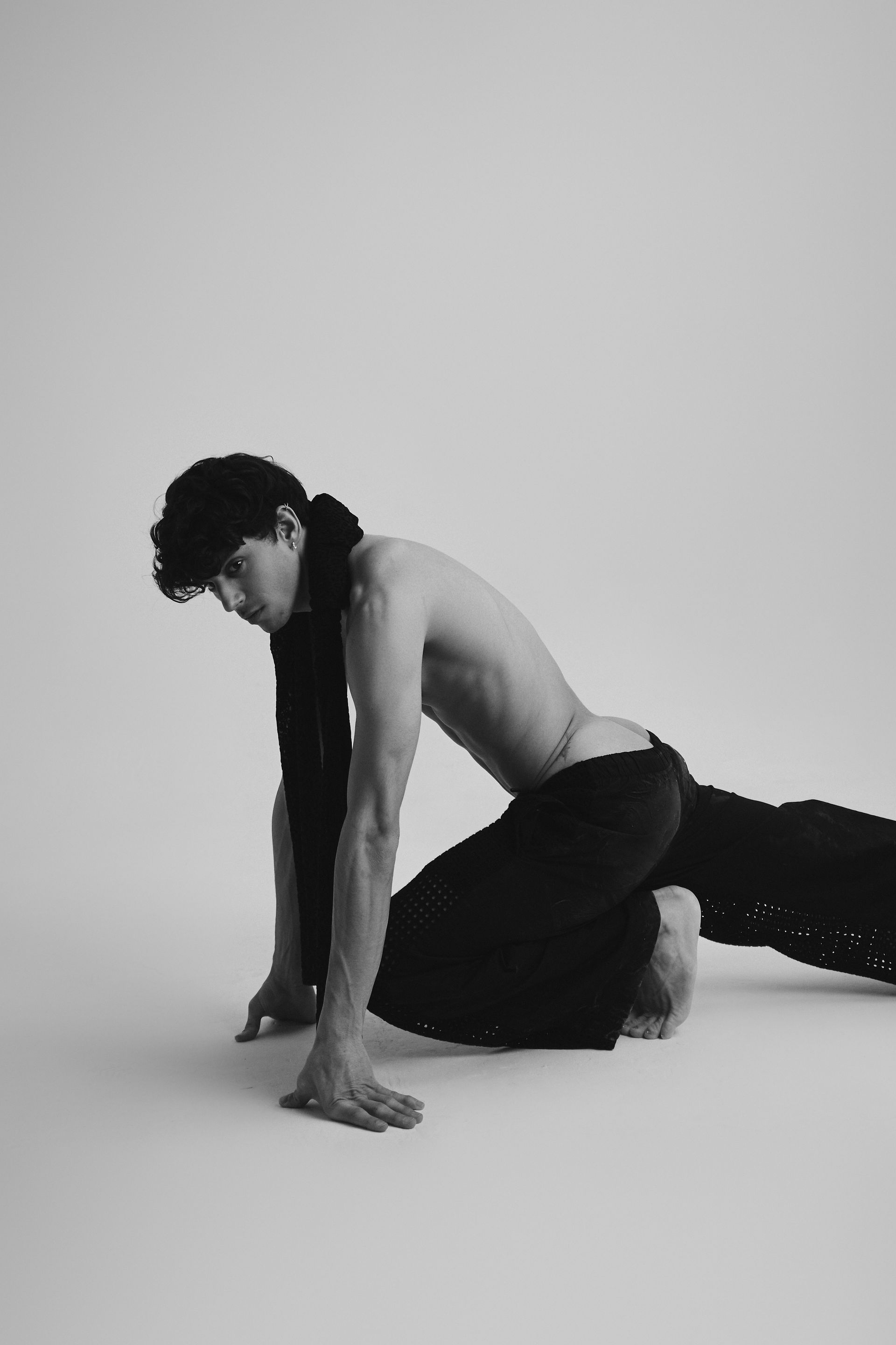 A person crouches on a white surface, wearing black pants and a dark scarf. Bare torso.