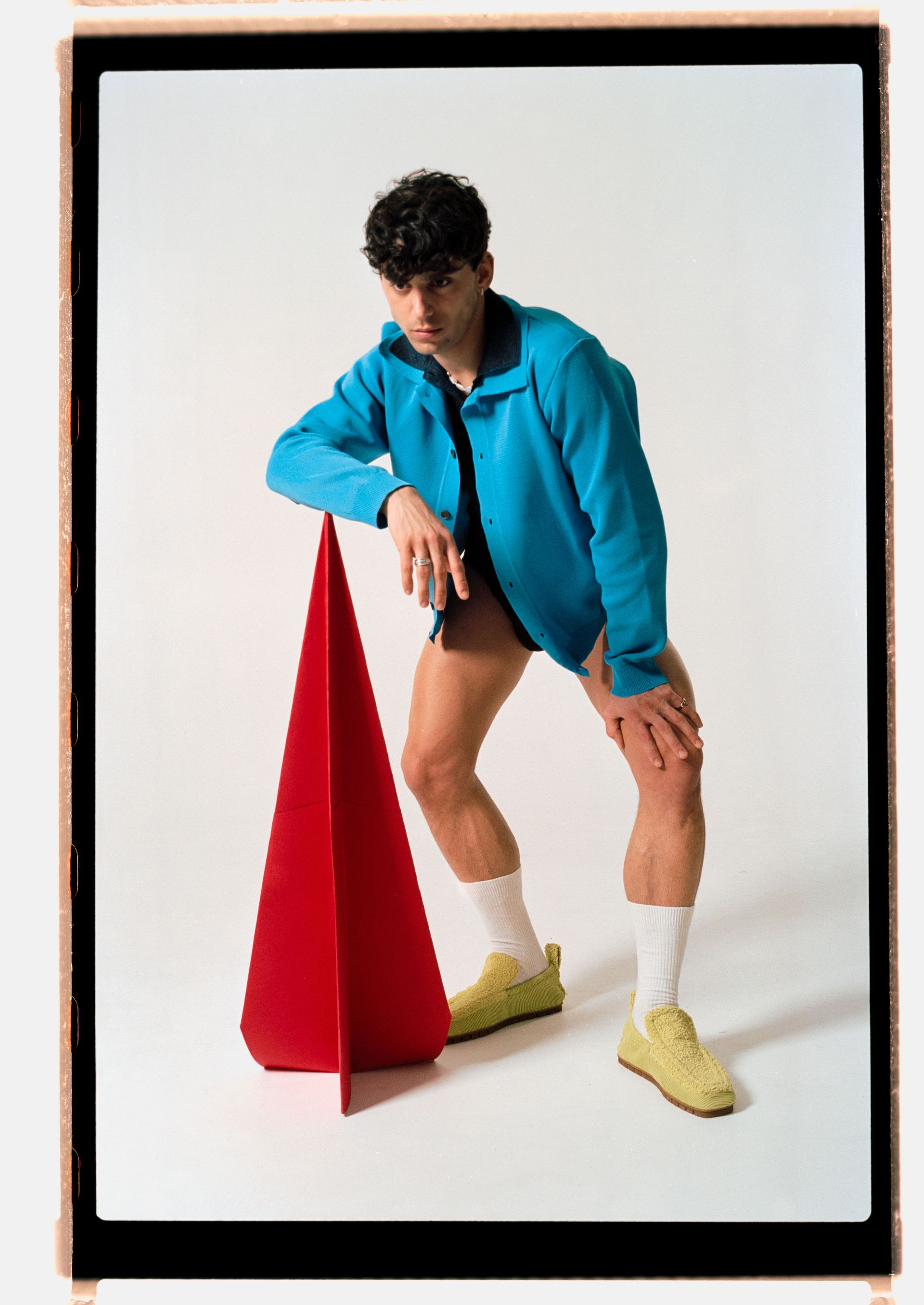 Person wearing blue jacket and shorts, leaning on red cone; white socks and yellow sneakers.
