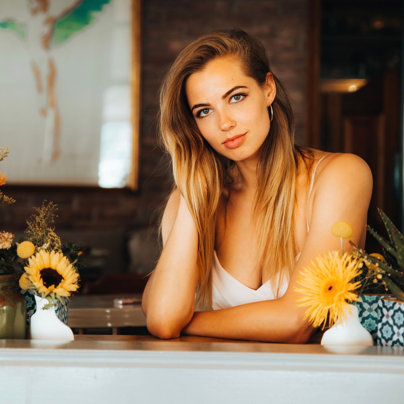 Blond woman leaning on a counter, surrounded by flowers, with a calm expression.