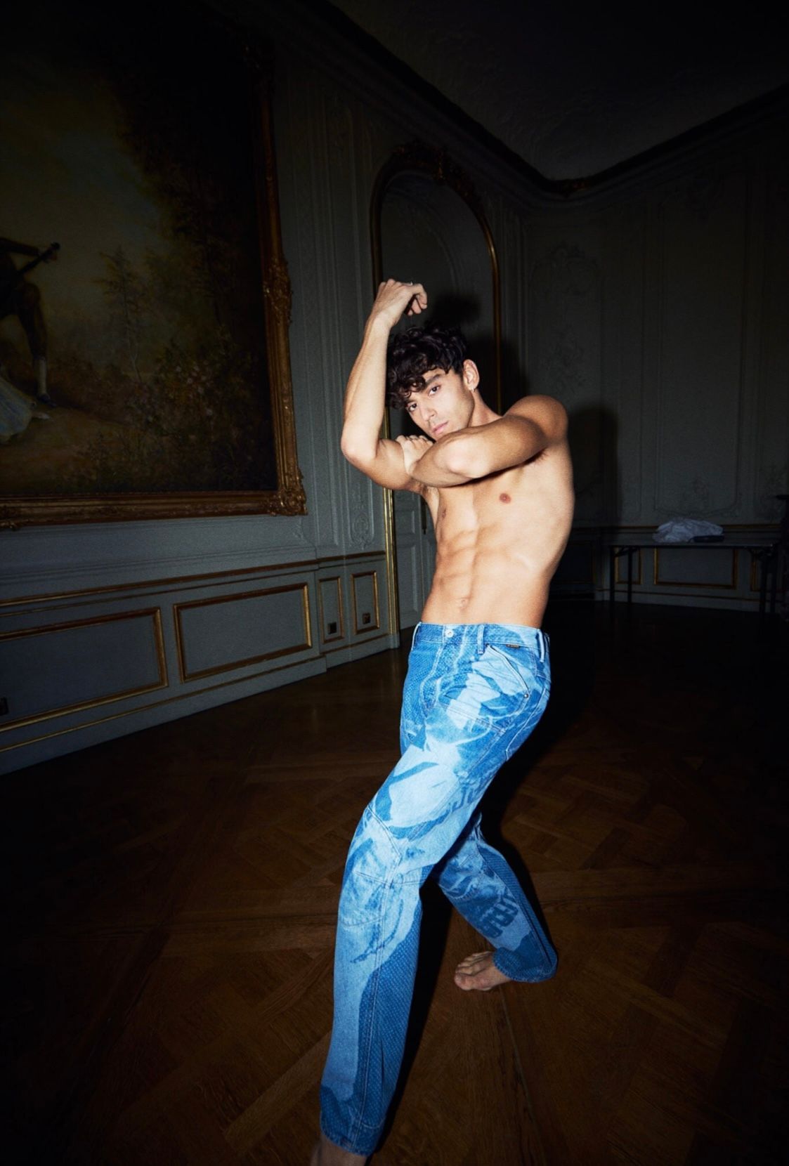 Bare-chested person in blue patterned jeans dances in a room with a painting and dark wood floor.