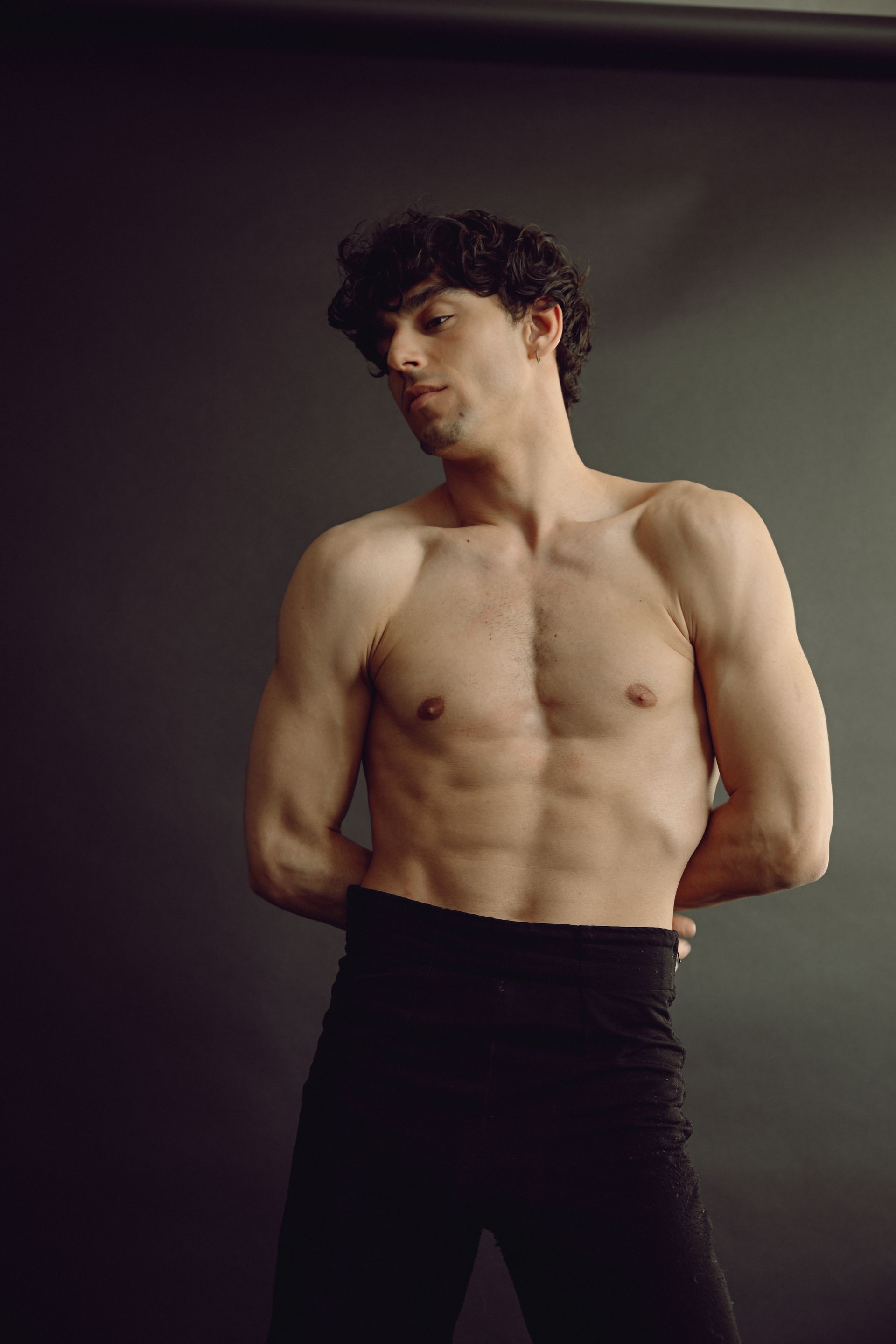 Man with curly hair, shirtless, hands behind back, wearing black pants, against a dark backdrop.