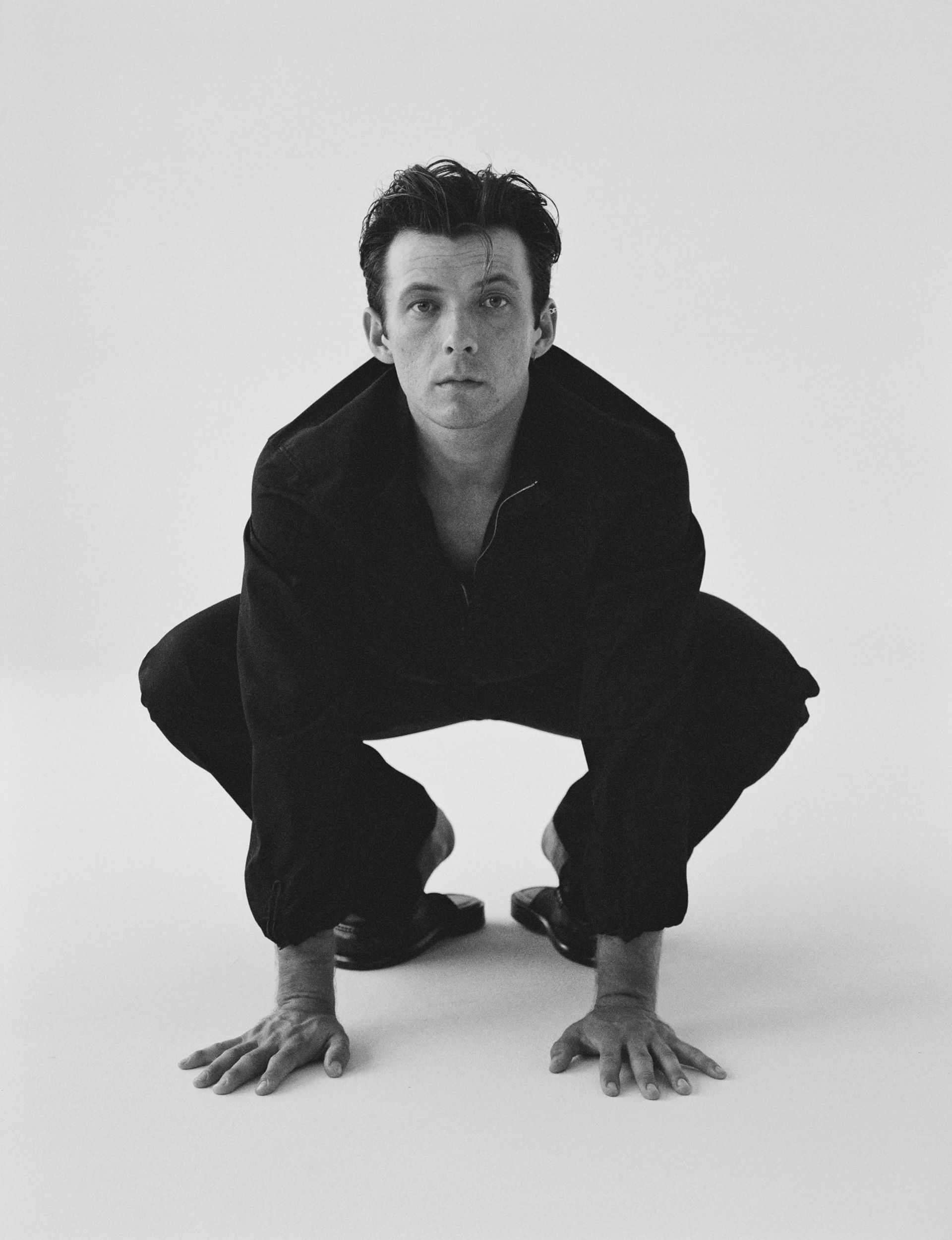 Man crouched low in black clothing, hands on the floor, looking directly at the viewer. White backdrop.