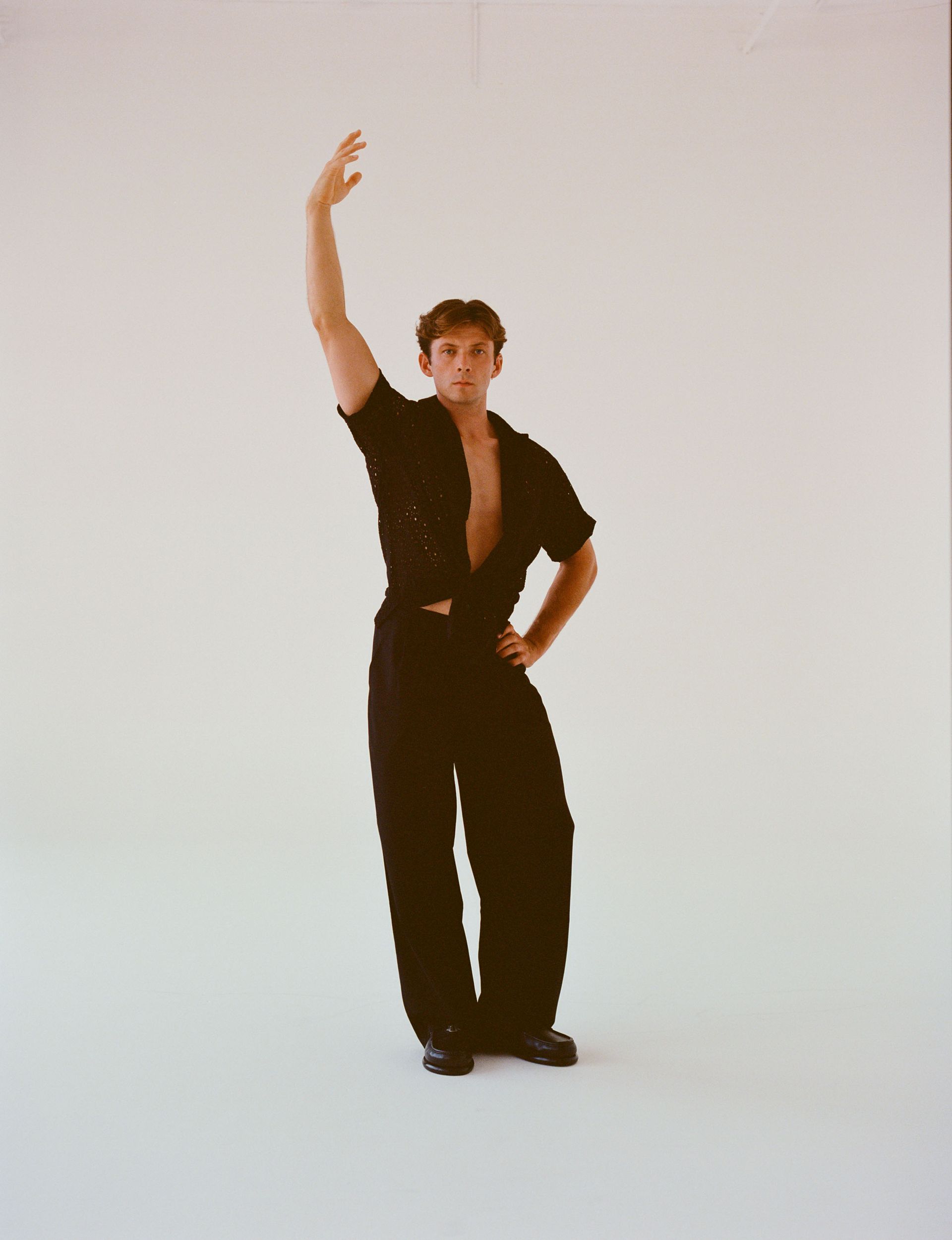 Person in black outfit, arm raised, hand on hip, against white background.
