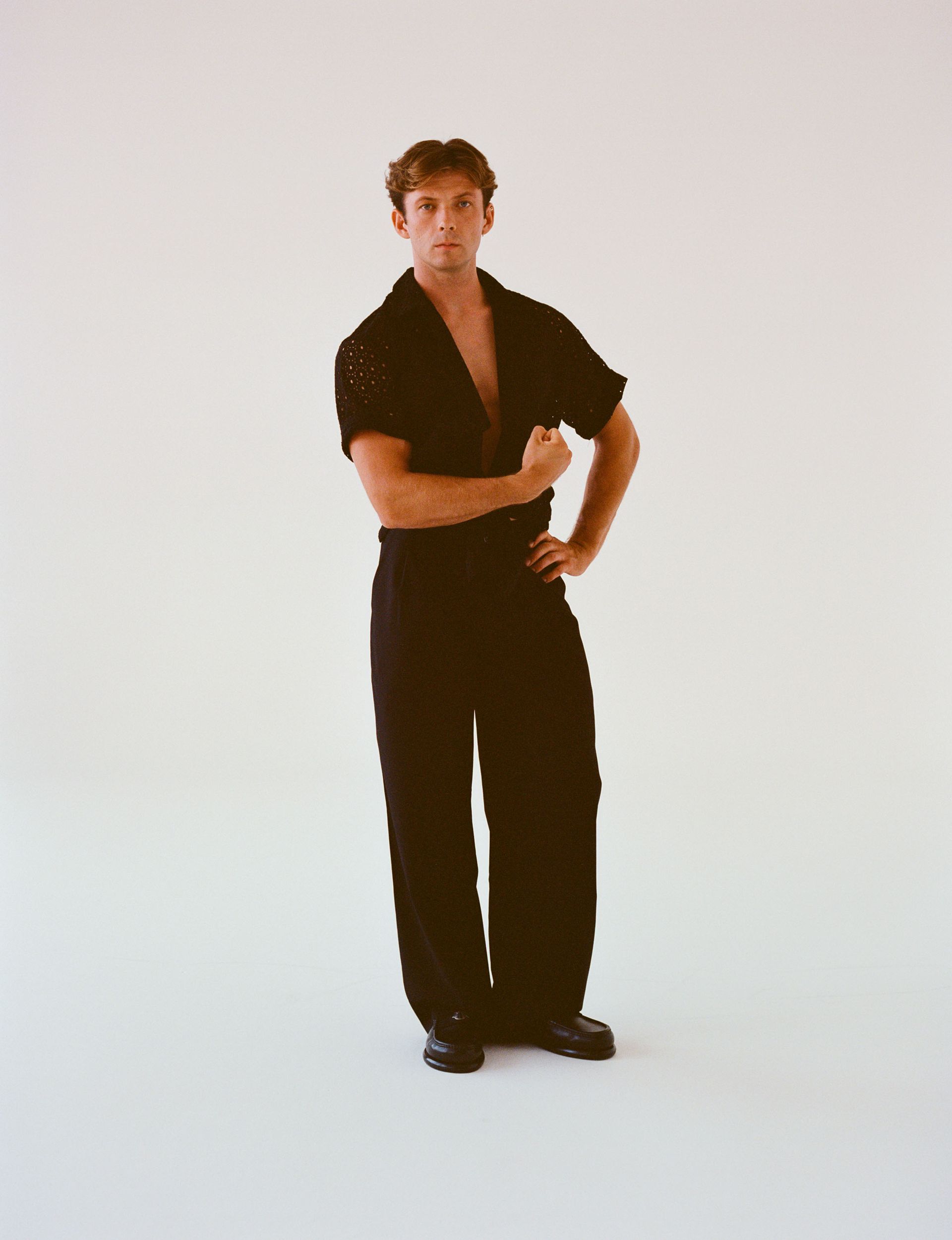 Man in black outfit, one arm flexed, hand on hip. Posing in front of a white background.