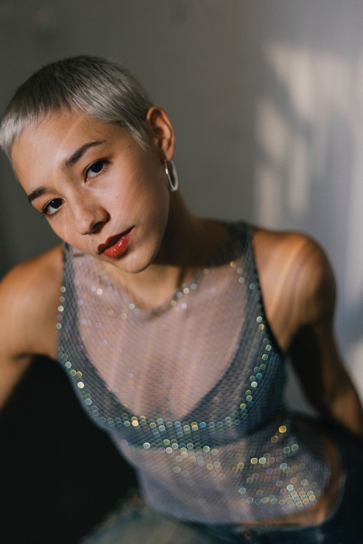 Woman with short, silver hair, wearing a sparkly mesh top. Leaning towards the camera.