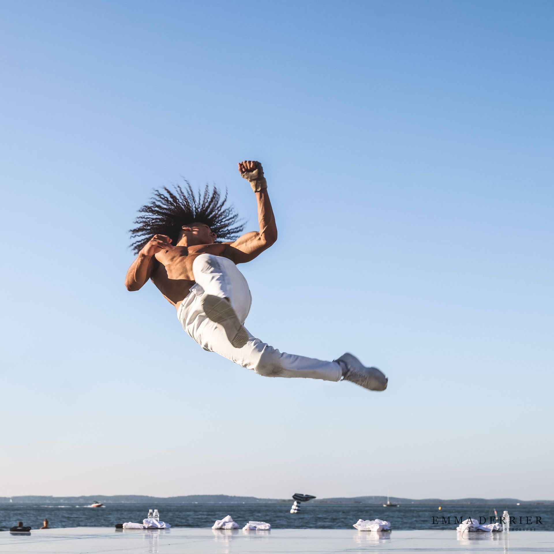 Man leaping in mid-air, fist raised, over water with boats, wearing white pants and sneakers; blue sky background.