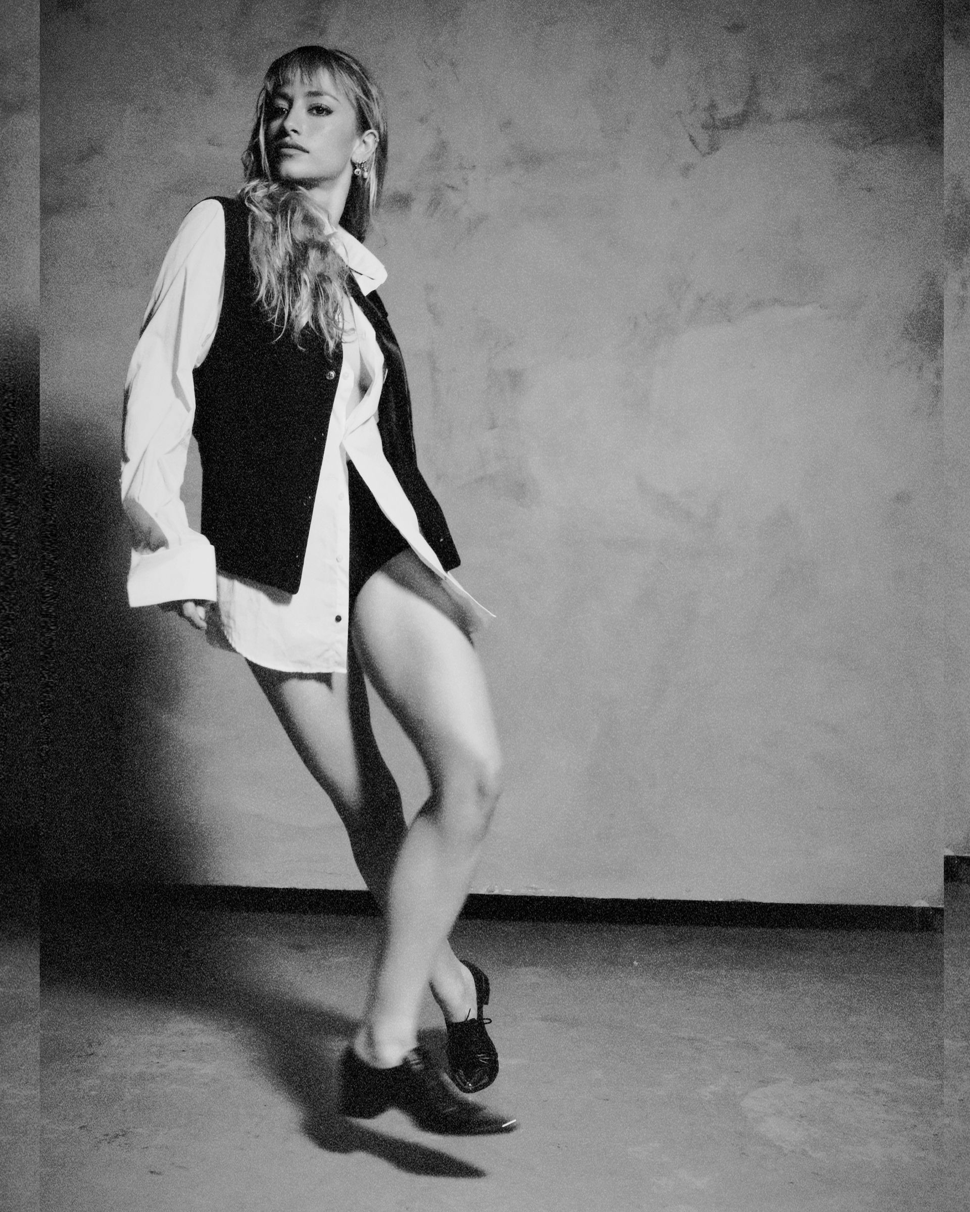 Woman in oversized white shirt and black vest, leaning against wall, wearing shoes.