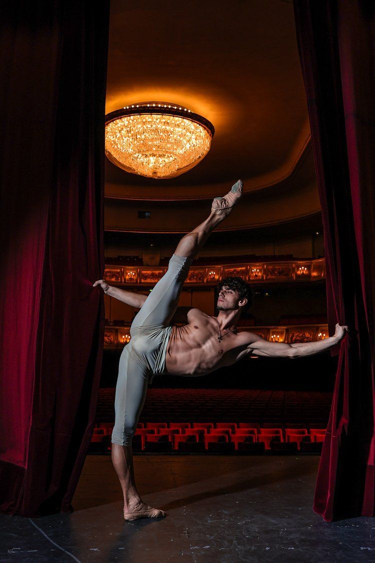 Male ballet dancer in a high split pose on stage, red curtains, gold chandelier, and empty audience seats.
