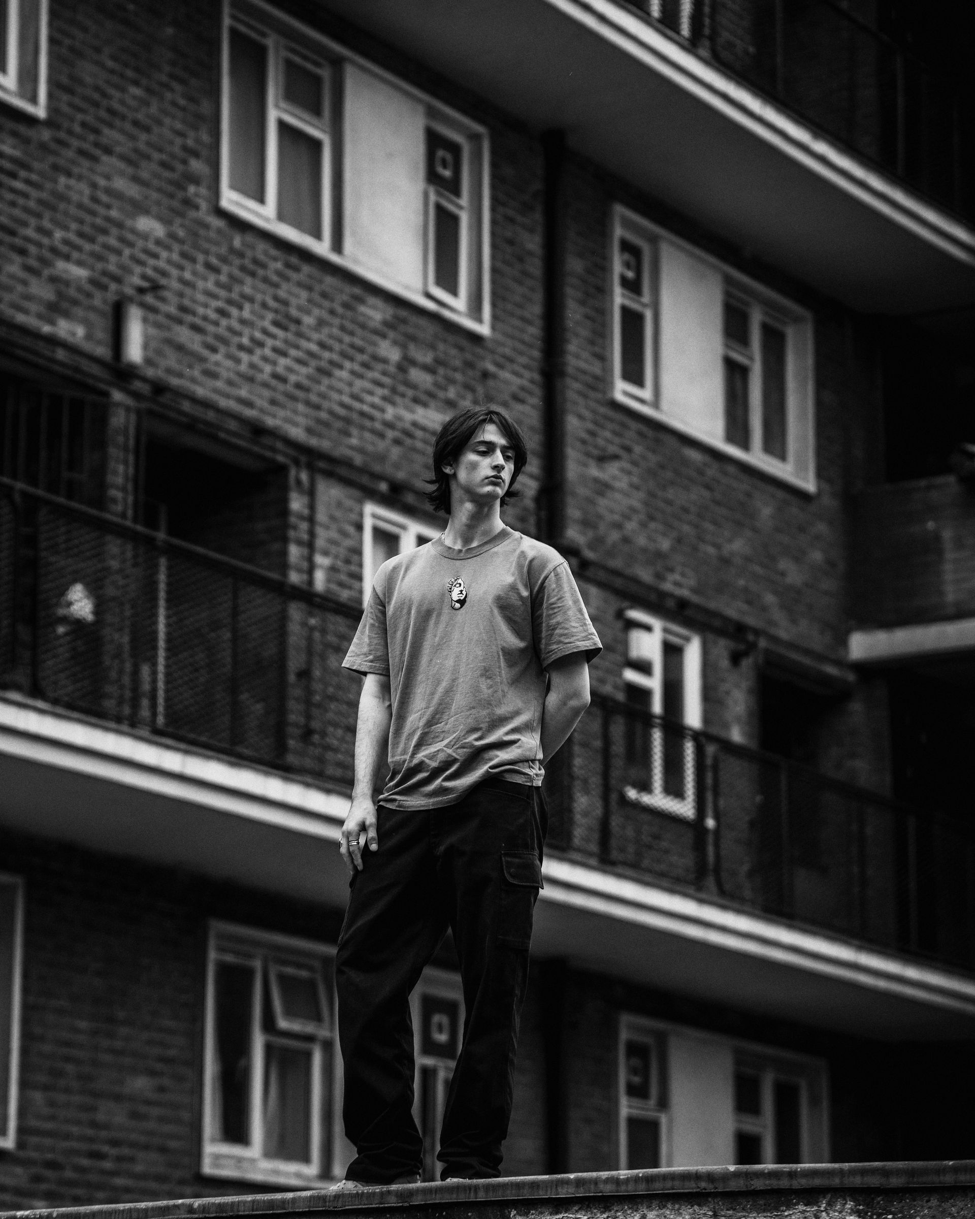 Man standing in front of a brick building with windows. He is wearing a t-shirt and dark pants.