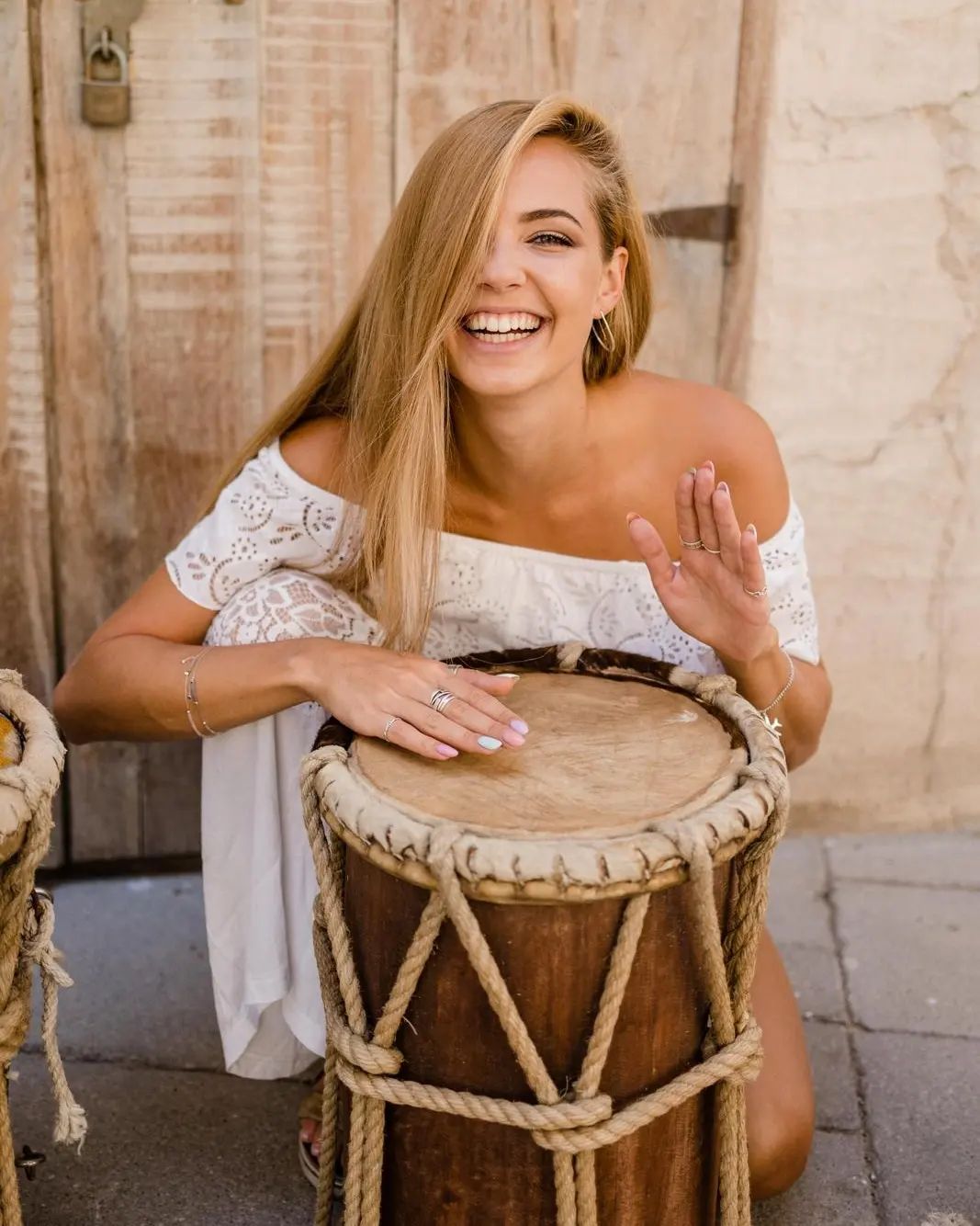 Woman smiling, playing a drum. She is wearing a white dress, outdoors.
