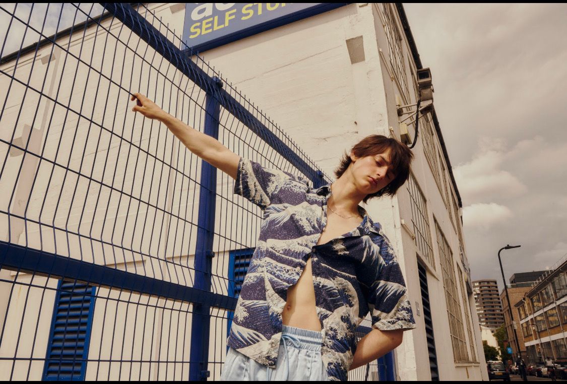 Man in open patterned shirt leans against a blue fence, pointing upwards near a building with 