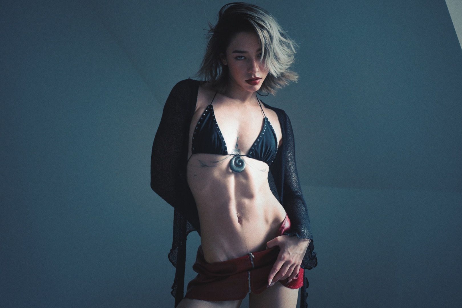 Woman in black bikini top and shorts, sheer black cover-up, hand on hip, in front of a blue background.