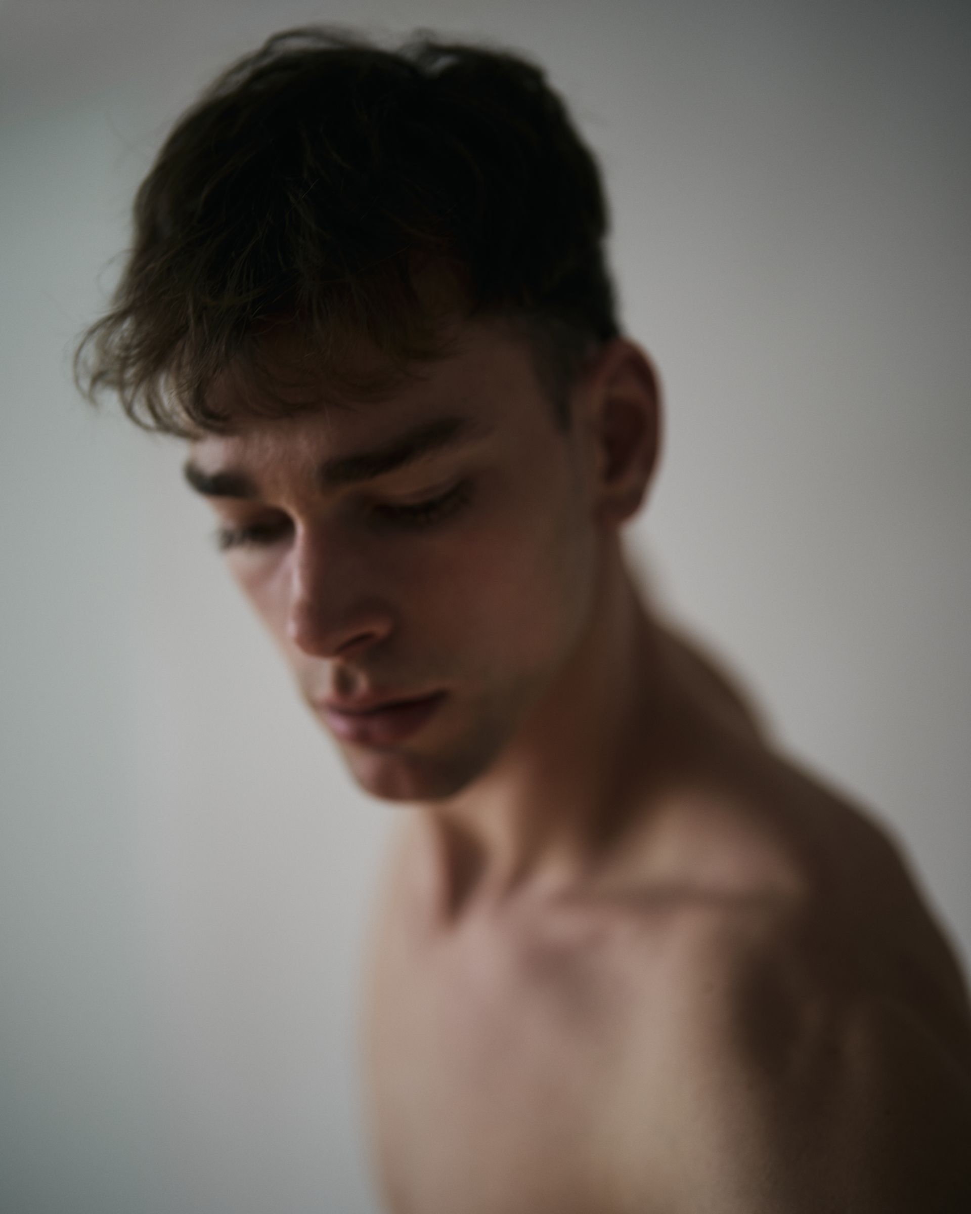 Man with dark hair and bare shoulders looking down against a neutral background.