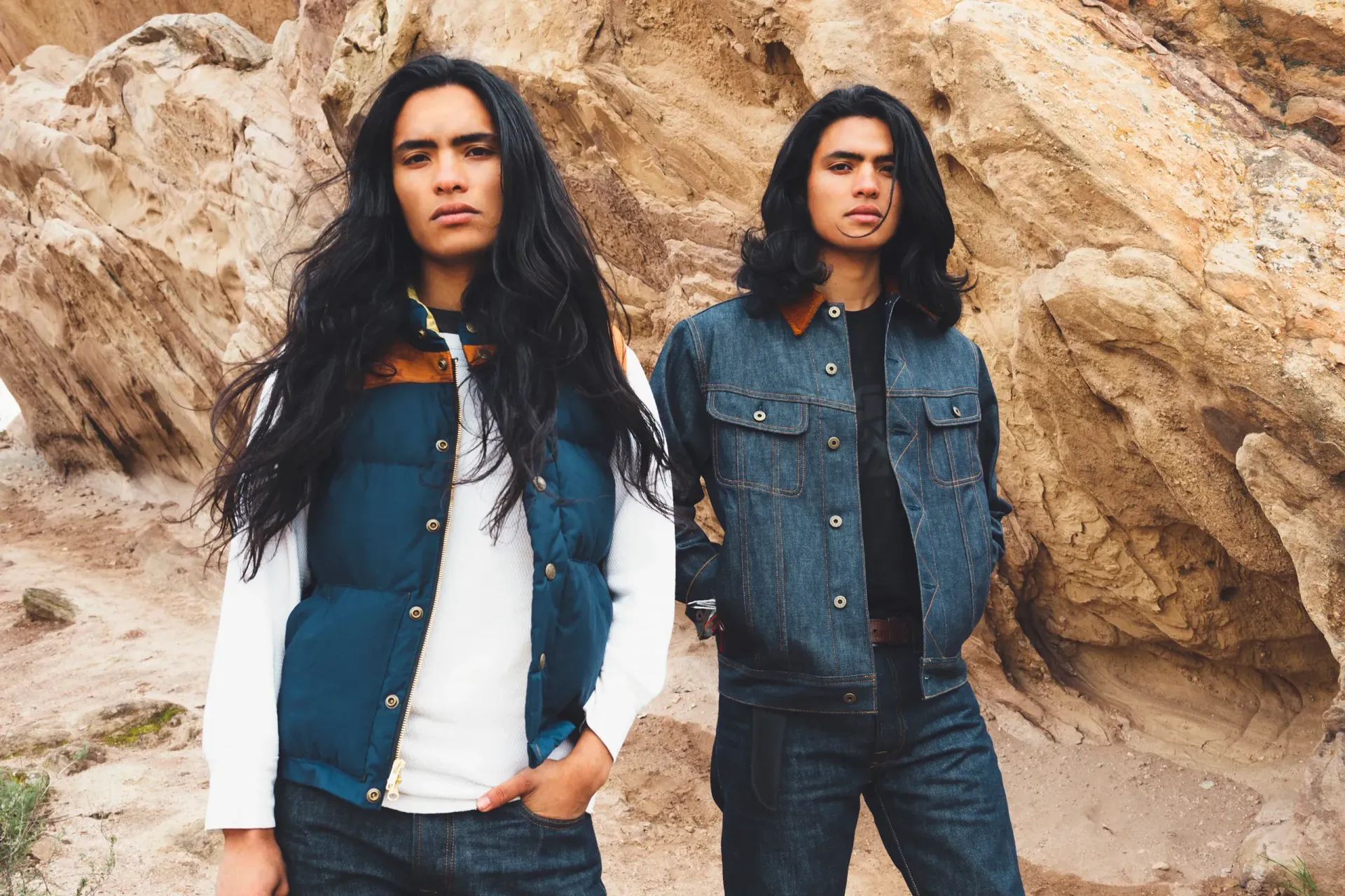 Two people with long, dark hair stand outside. One wears a blue vest, the other a denim jacket. They face forward.