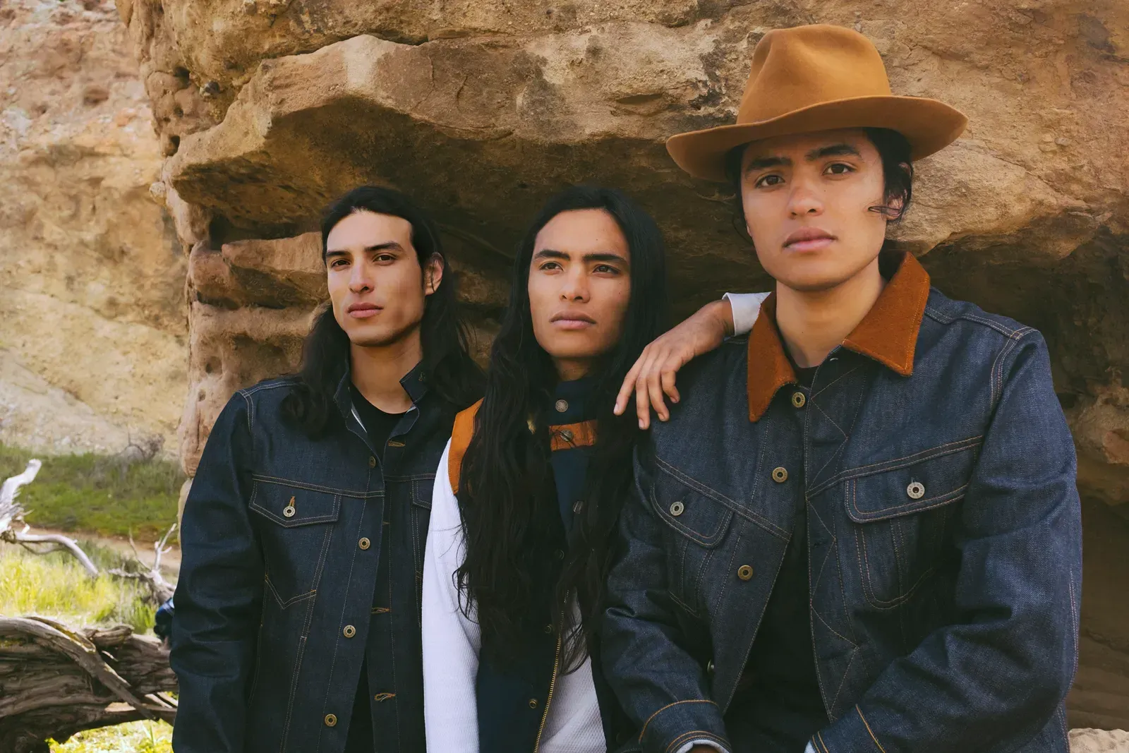 Three people in denim jackets and a brown hat against a rock formation, looking towards the camera.