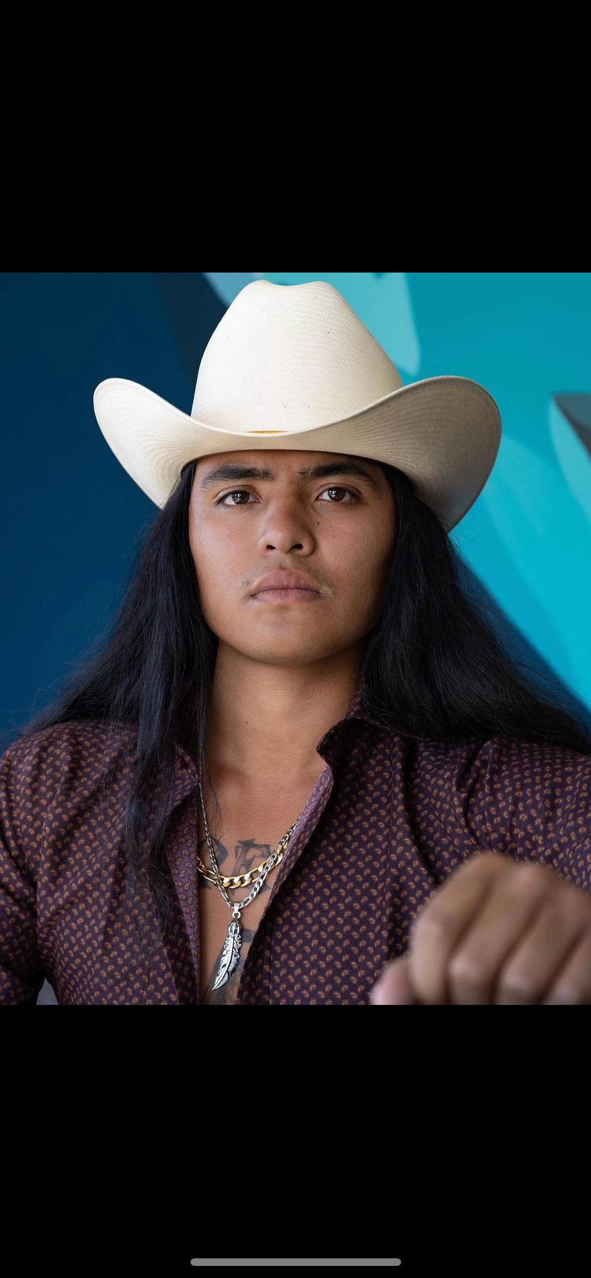 A person wearing a cowboy hat, long hair, and a dark shirt with a necklace, against a blue background.