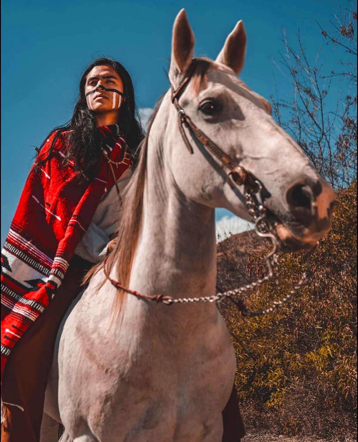 Person on a white horse, wearing red patterned shawl. Blue sky, dry brush in background.