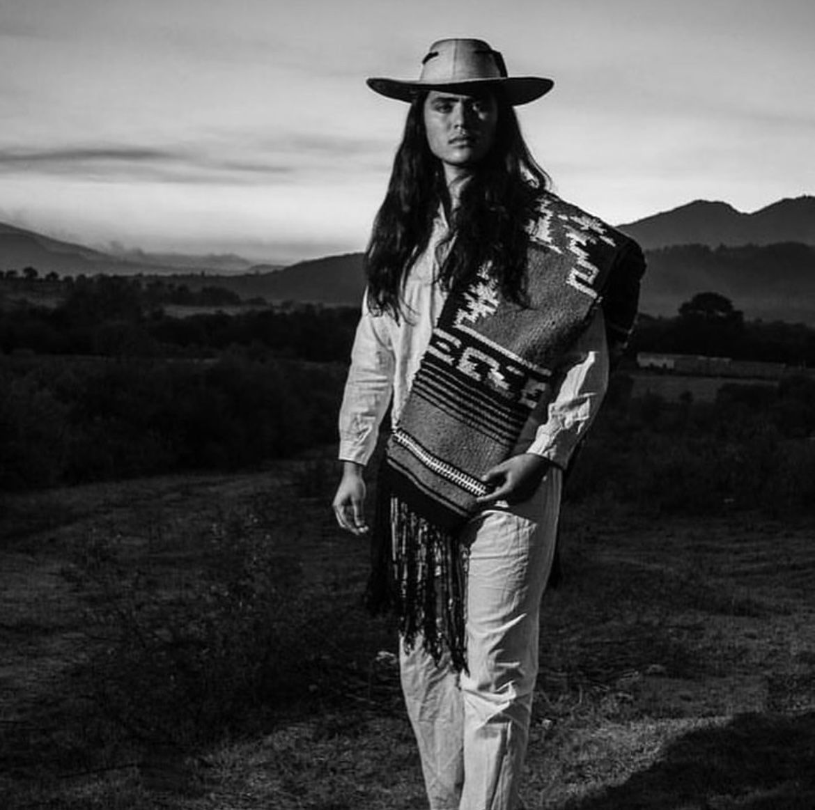 Woman wearing a hat and shawl in a field at sunset, holding the shawl in front of her. Mountains in the background.