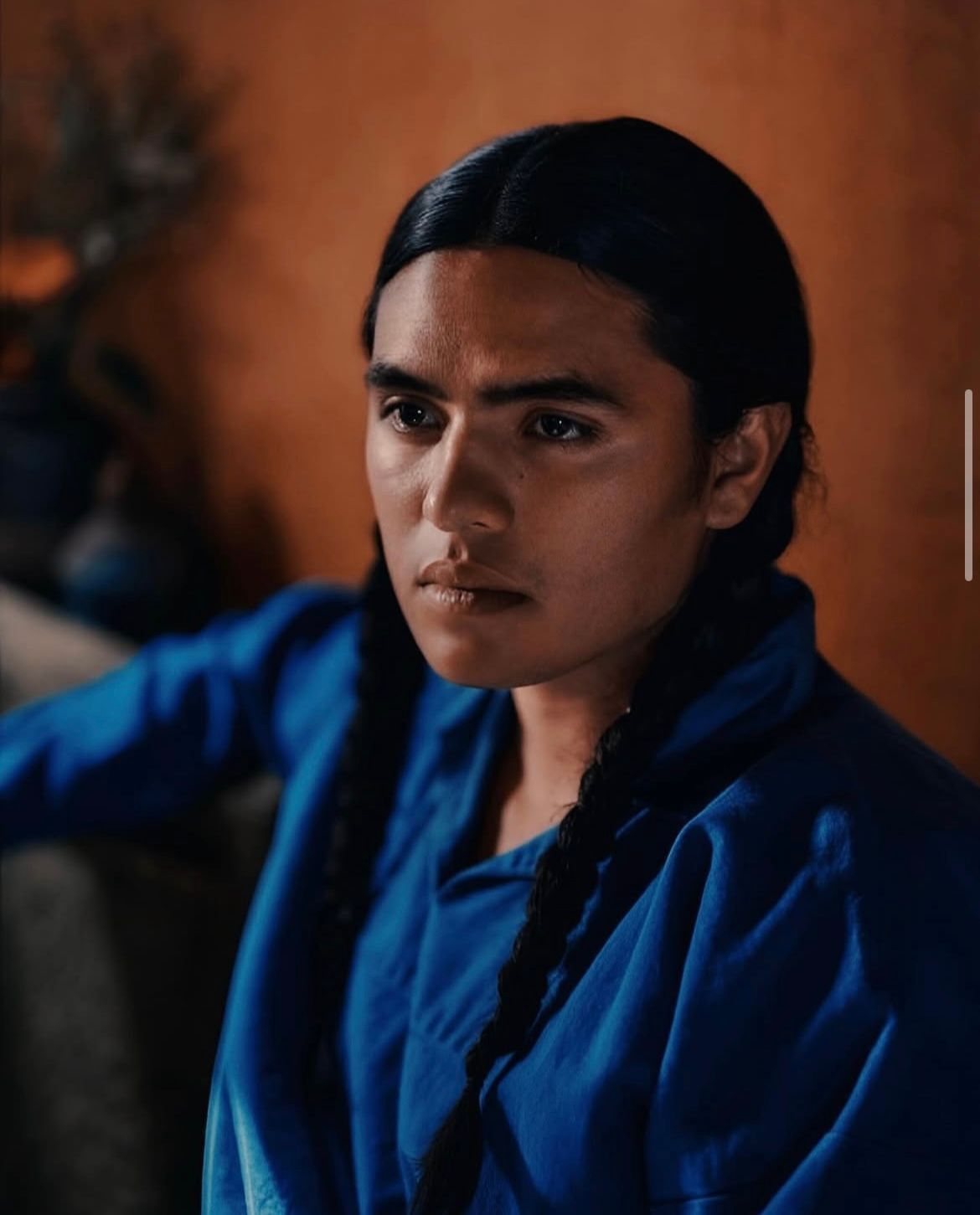 Person with dark hair in blue shirt, braided, looking away. Warm-toned background.