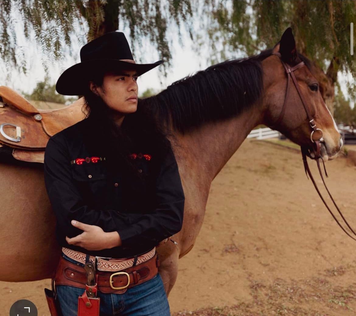 Cowboy in black shirt and hat, leaning on brown horse in a dusty outdoor setting.