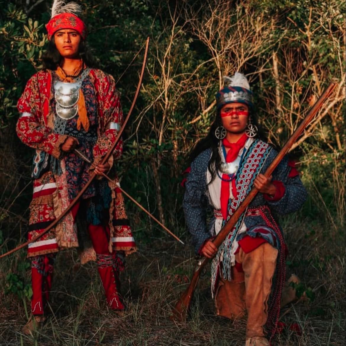 Two people in embellished traditional attire holding bows in a grassy outdoor setting.