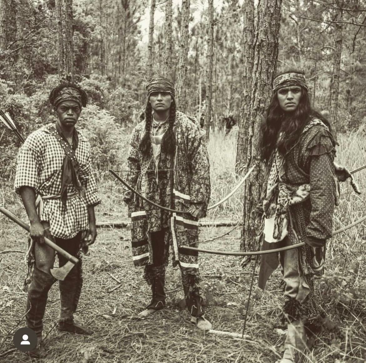 Three people in wooded area, dressed in period clothing, with bows and an axe.