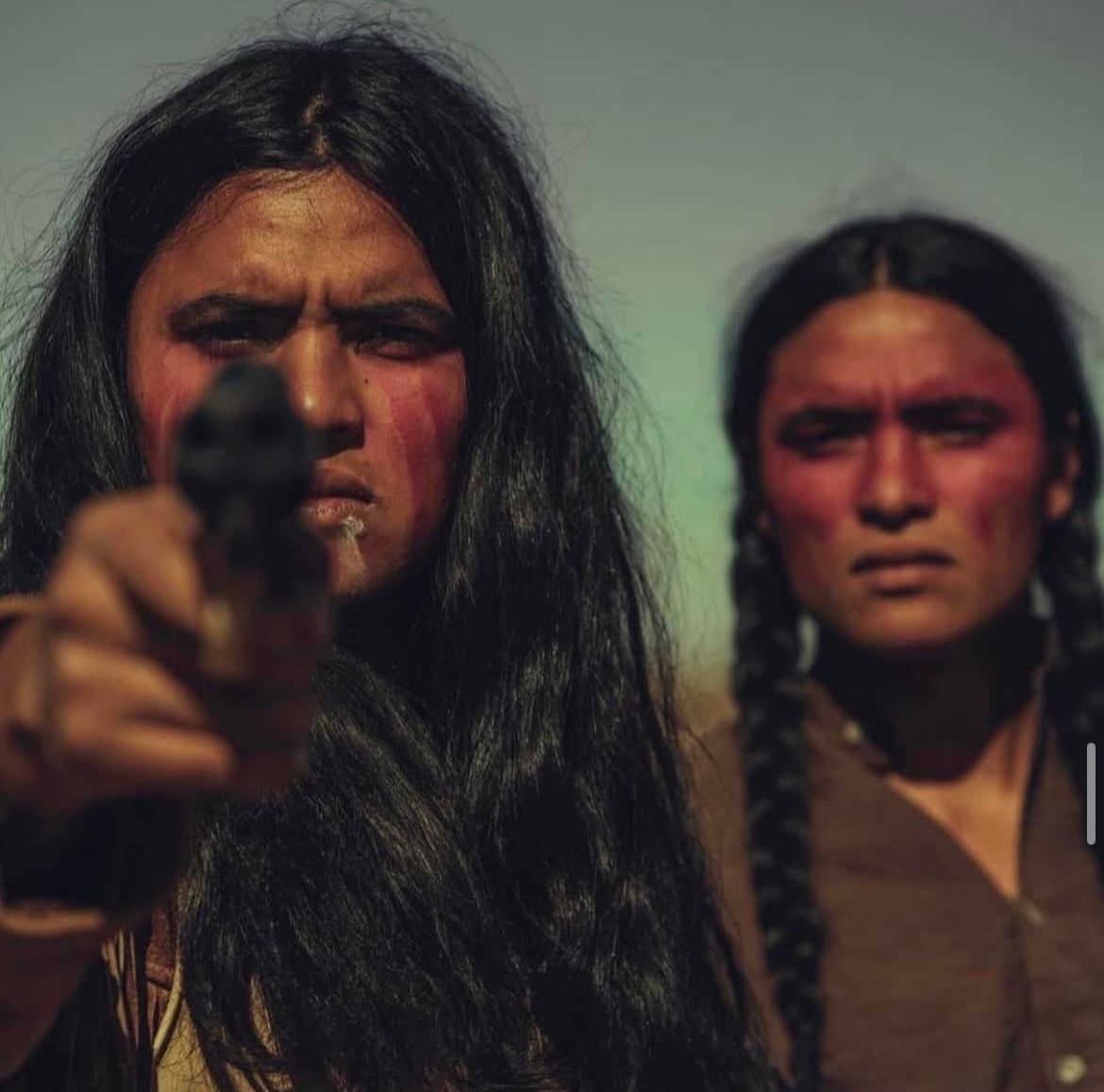 Two people, one holding a pistol, face the viewer with serious expressions. Red paint on their faces.