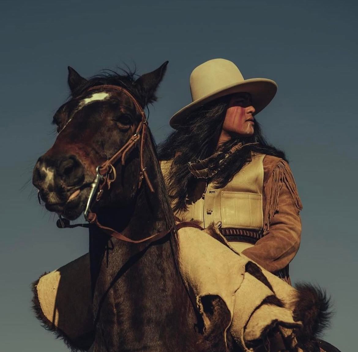 Person in cowboy hat rides a dark horse; dressed in buckskin, against blue sky.