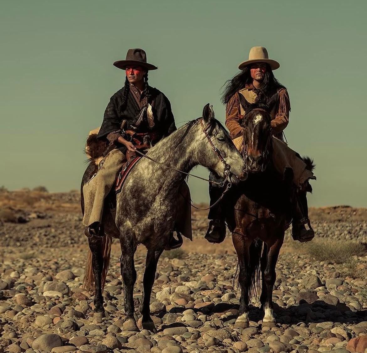 Two people on horseback in desert landscape; one on gray horse, one on brown.