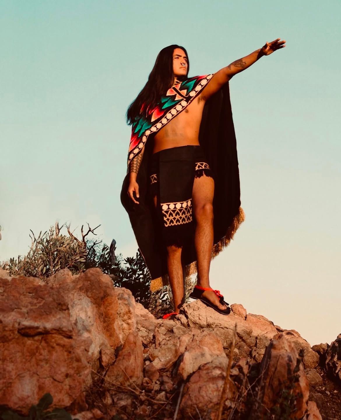 Man standing on a rock, arm outstretched, wearing a patterned shawl and skirt, under a blue sky.