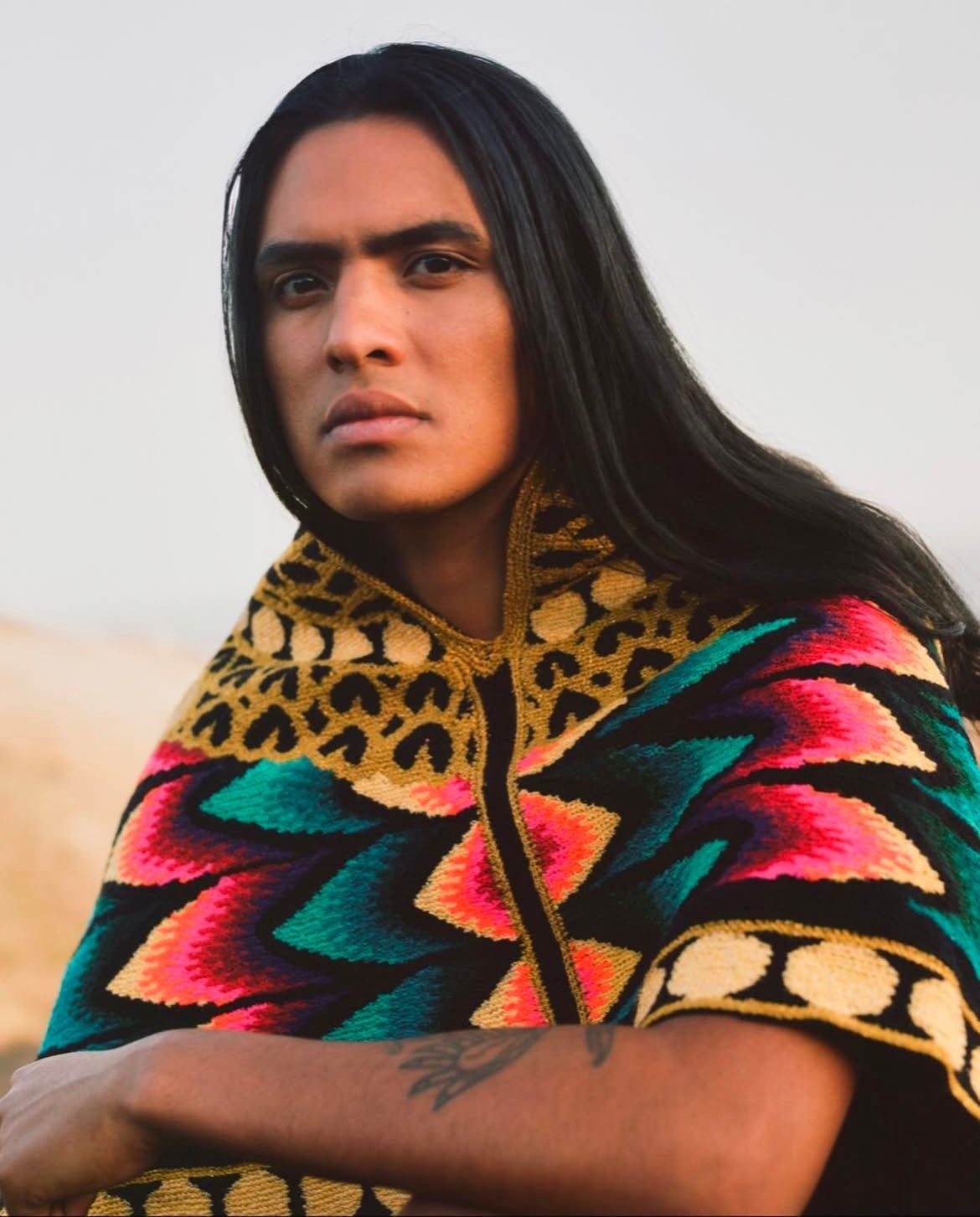 Man with long dark hair, wearing a colorful patterned shawl, looking at the camera.