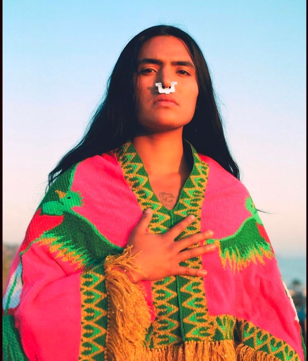Person with long dark hair, wearing a pink and green robe, hand on chest, nose ring.